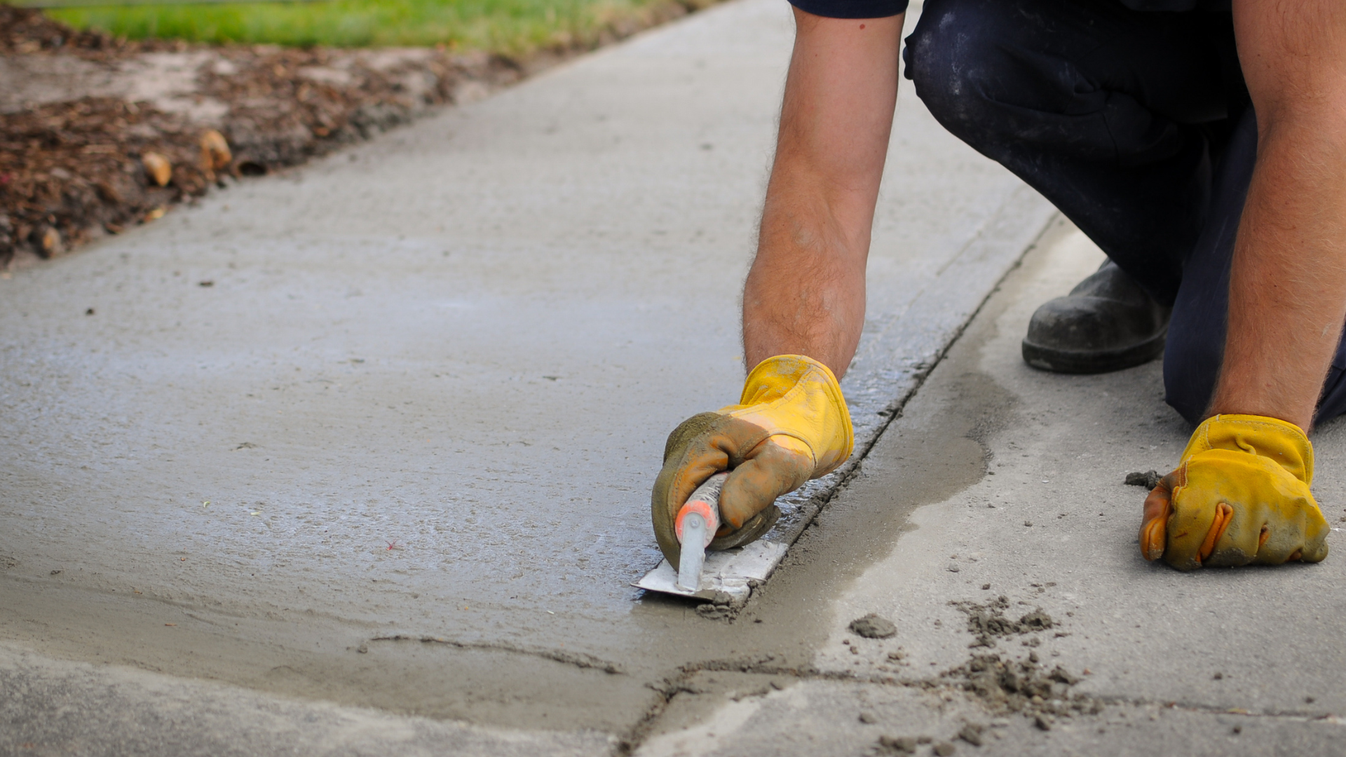 Worker smoothing wet concrete on a sidewalk with a trowel, wearing yellow gloves.