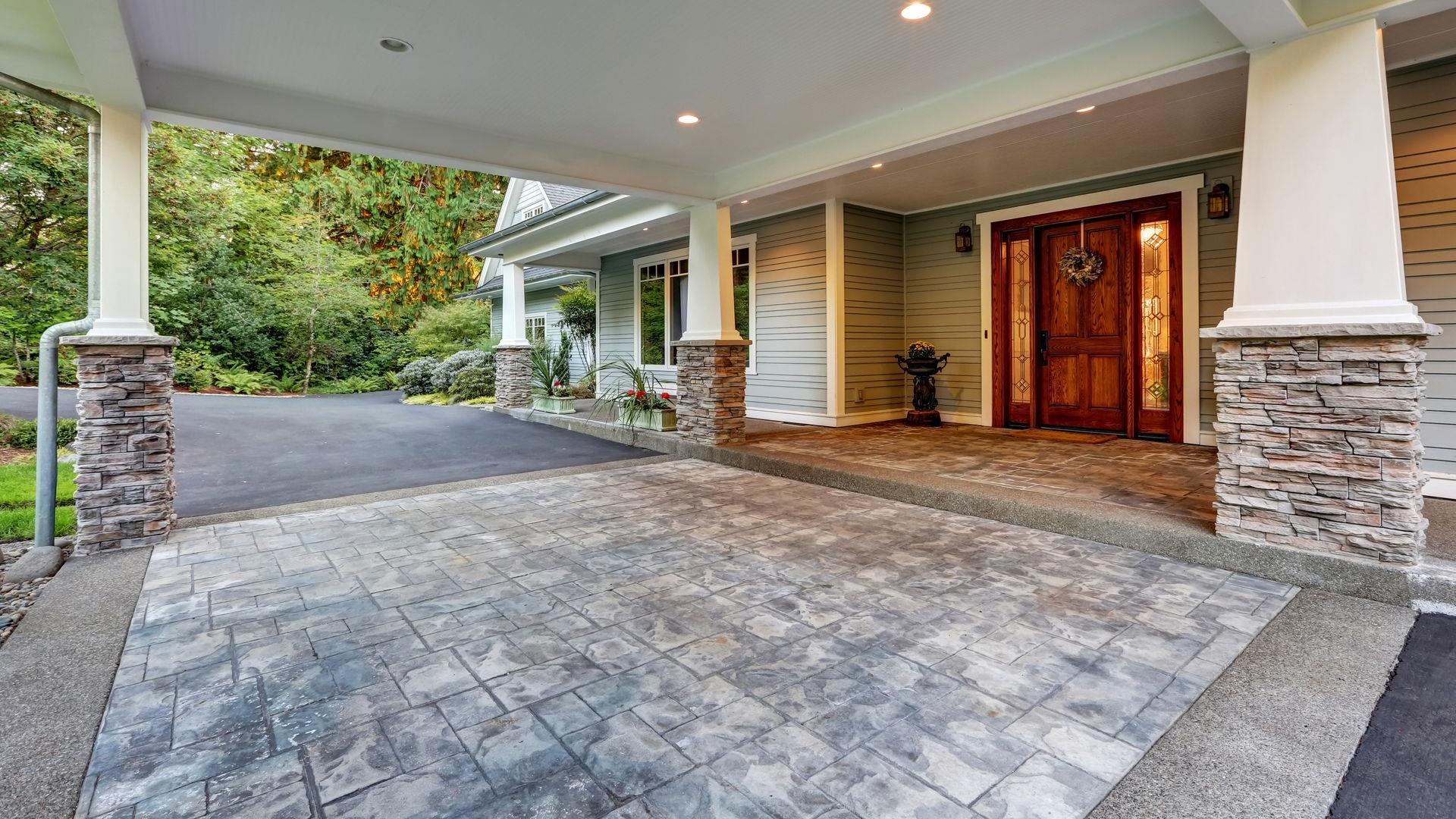 Covered stone entryway with wooden front doors and driveway leading to a house