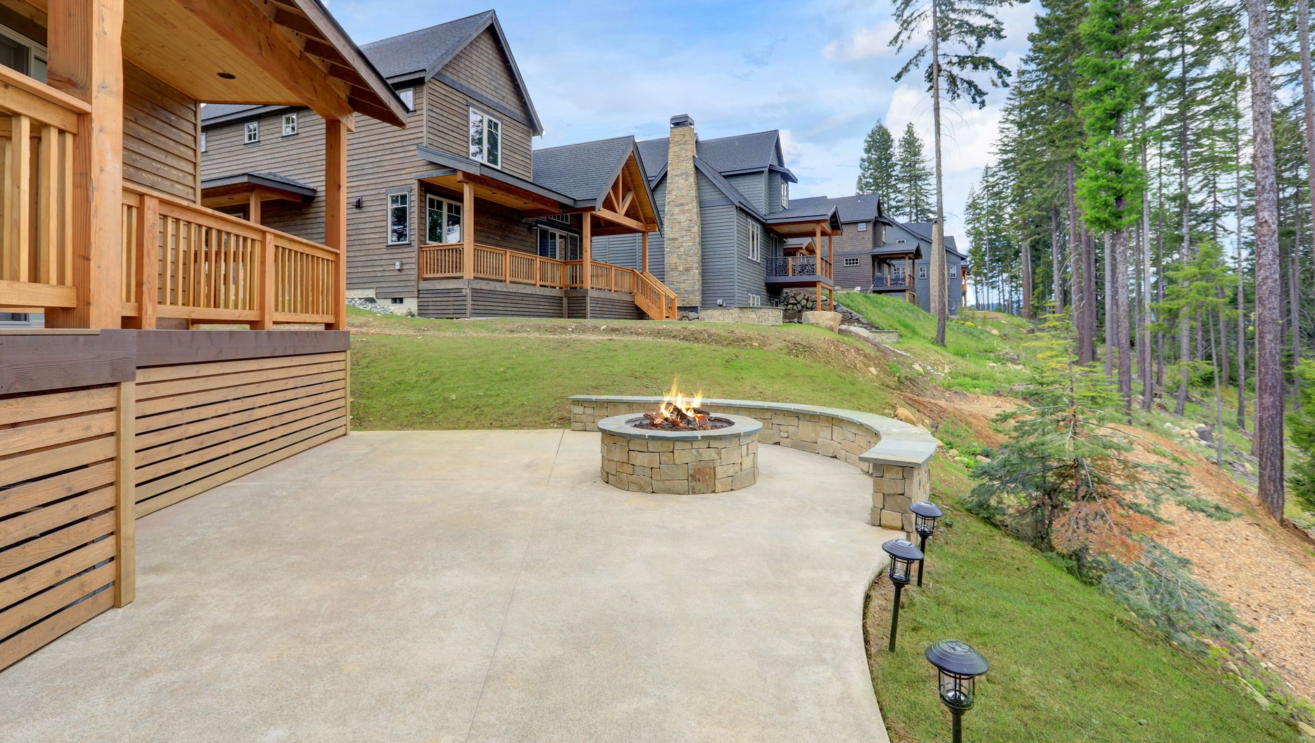 Stone patio with fire pit beside a rustic lodge and pine trees on a hillside