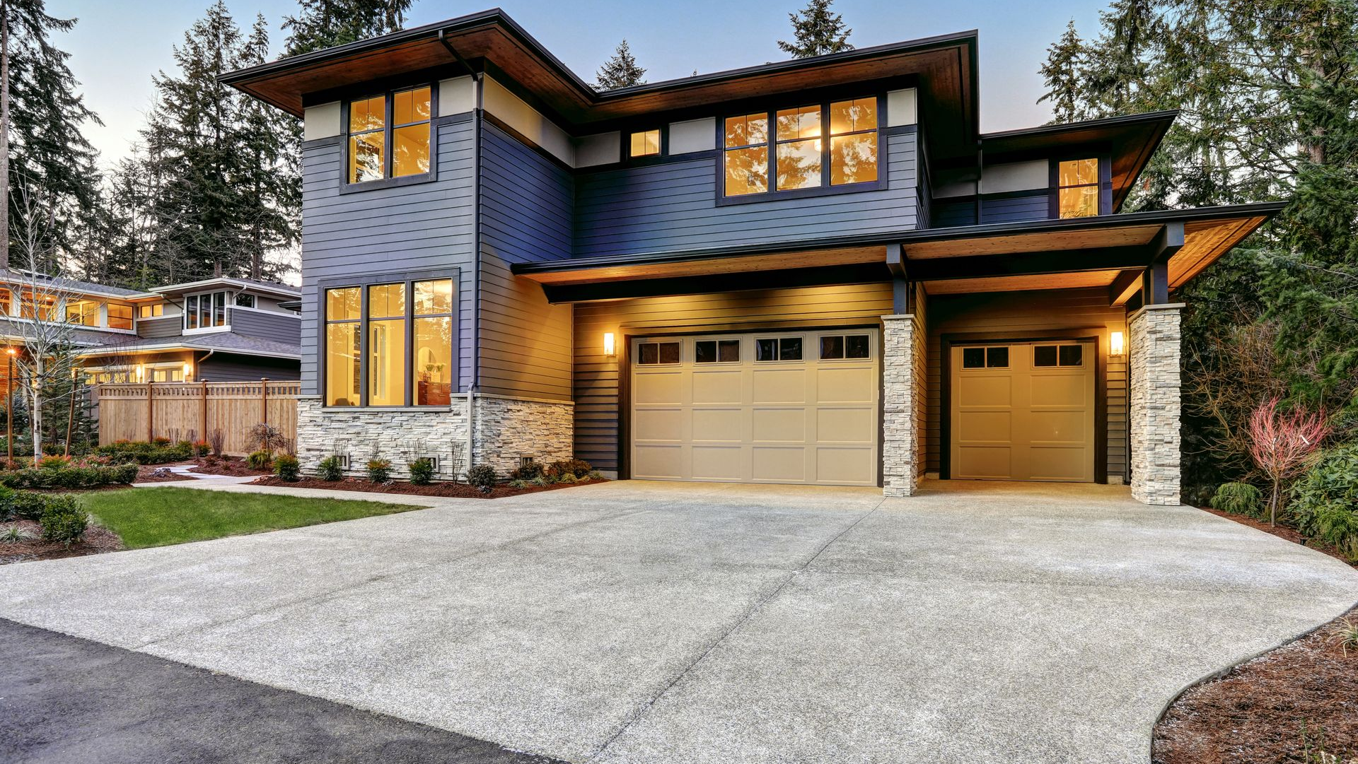 Modern two-story gray home with warm-lit windows, double garage, and a large concrete driveway at dusk
