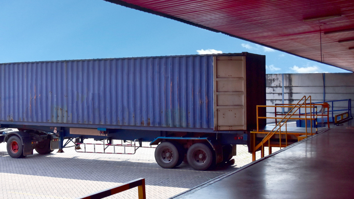 Semi-trailer truck parked at a loading dock under a red roof, viewed from the side.
