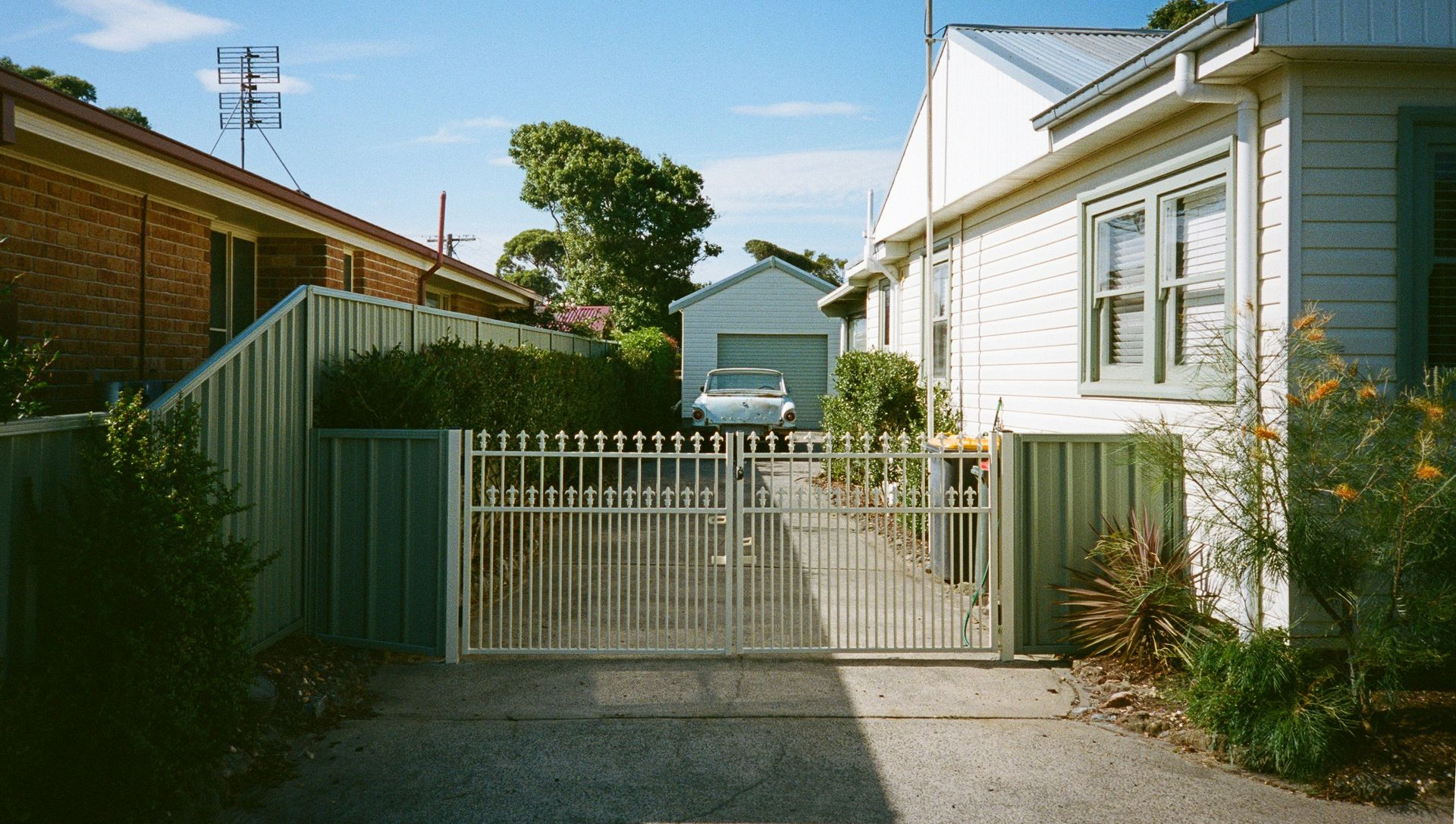 Driveway leading to a white house with a metal gate and a garage in the background. Blue sky.