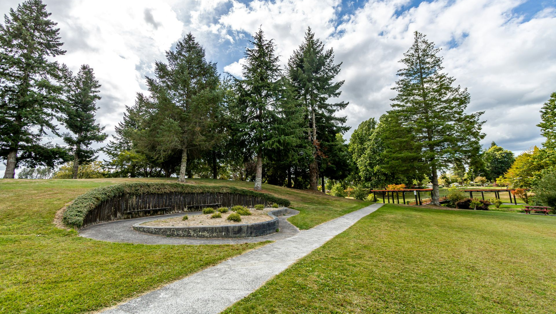 Stone monument with engraved plaque in a grassy park, bordered by trees and a gravel path under a cloudy sky