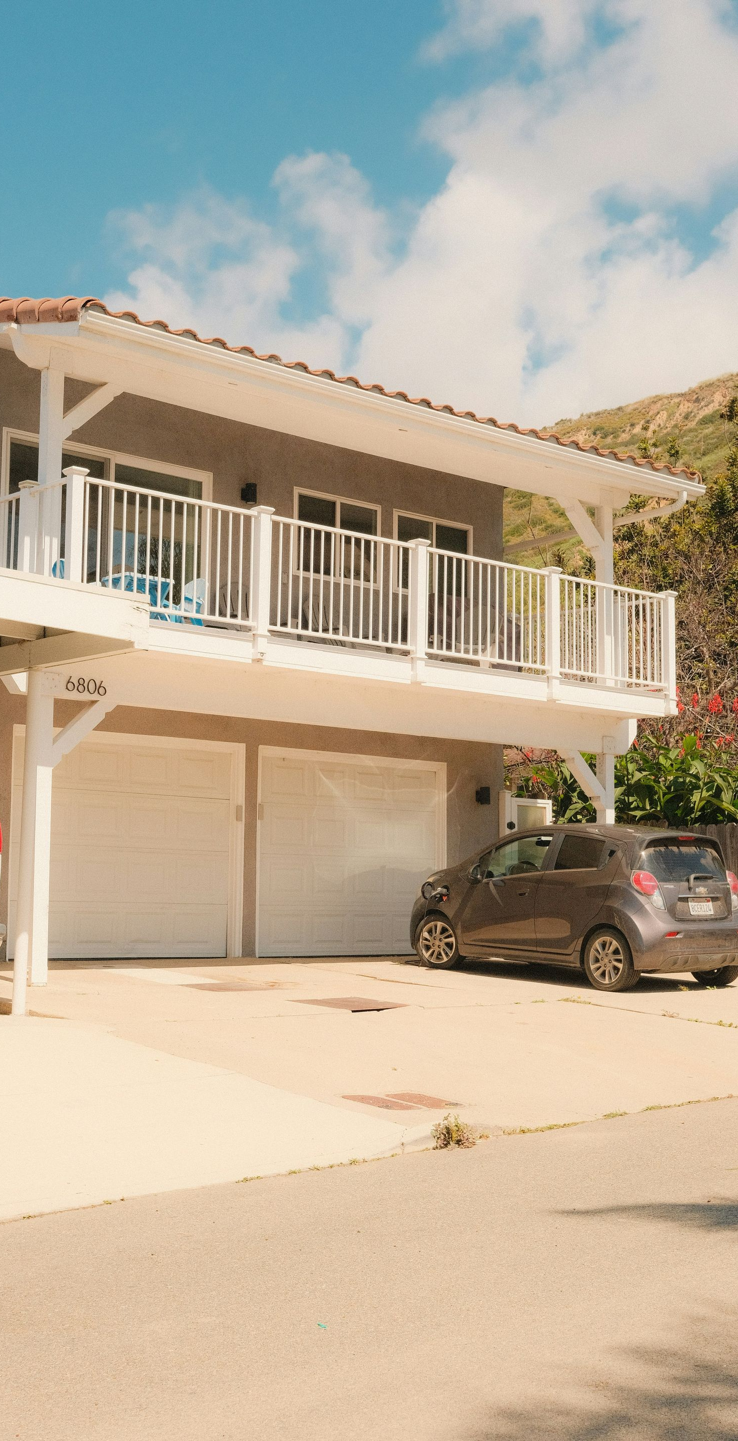 Driveway with parked cars beside a white two-story house and balcony under a blue sky