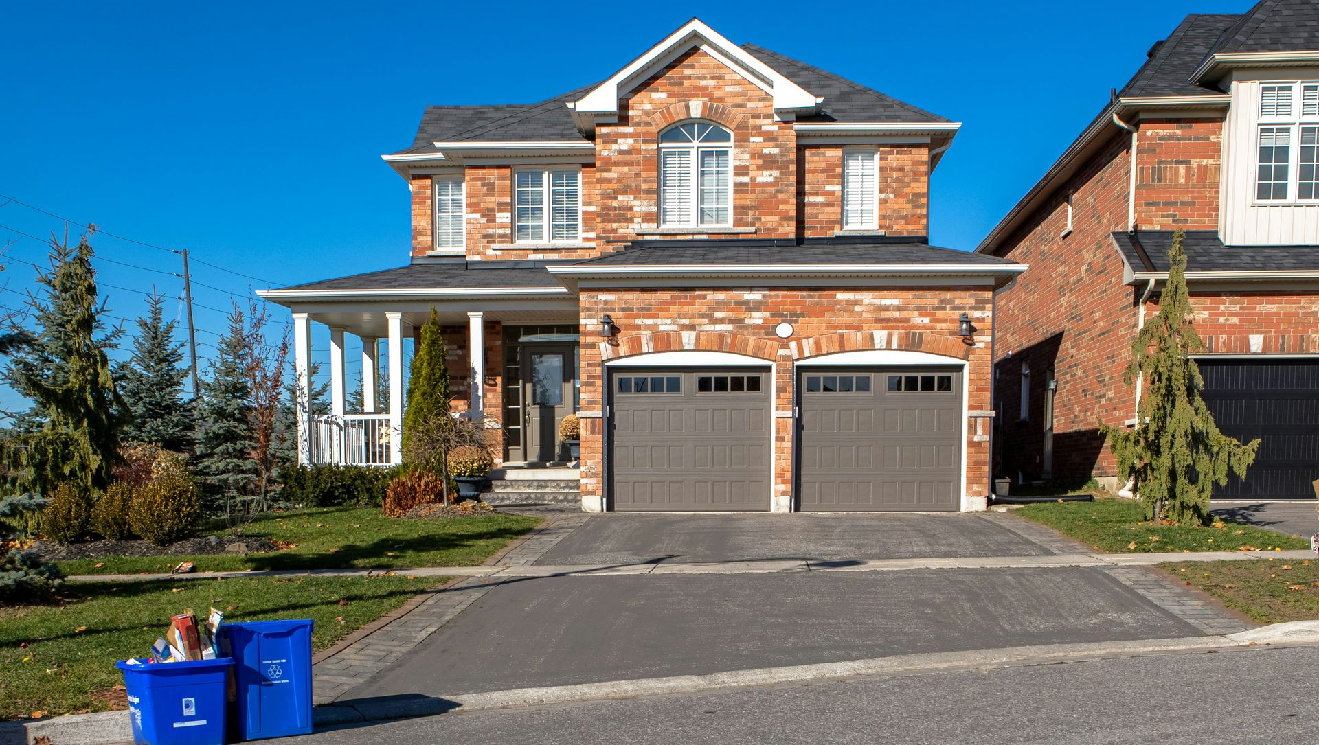 Two-story brick house with a gray garage door and blue recycling bins in front of it. Clear blue sky.