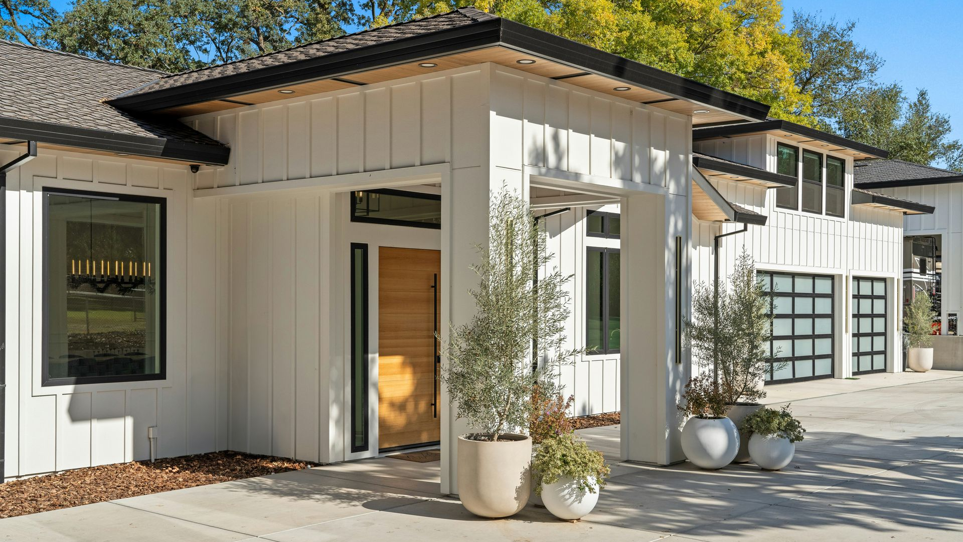 Modern white farmhouse with vertical siding, a wooden front door, glass garage doors, and potted plants on a patio.