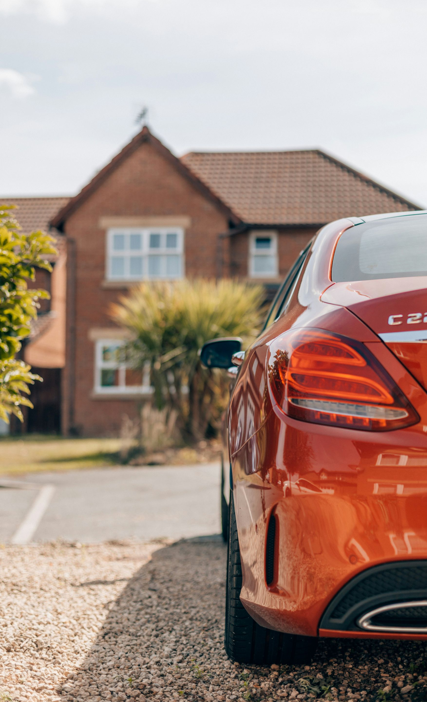 Orange Mercedes-Benz C220 parked on gravel in front of a brick house on a sunny day.
