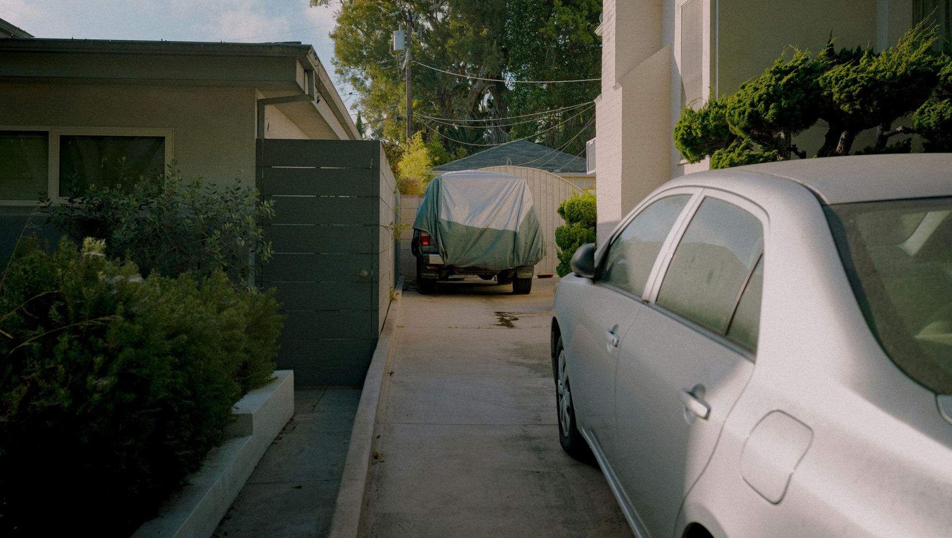 A car parked in a driveway next to a covered truck, between two houses.
