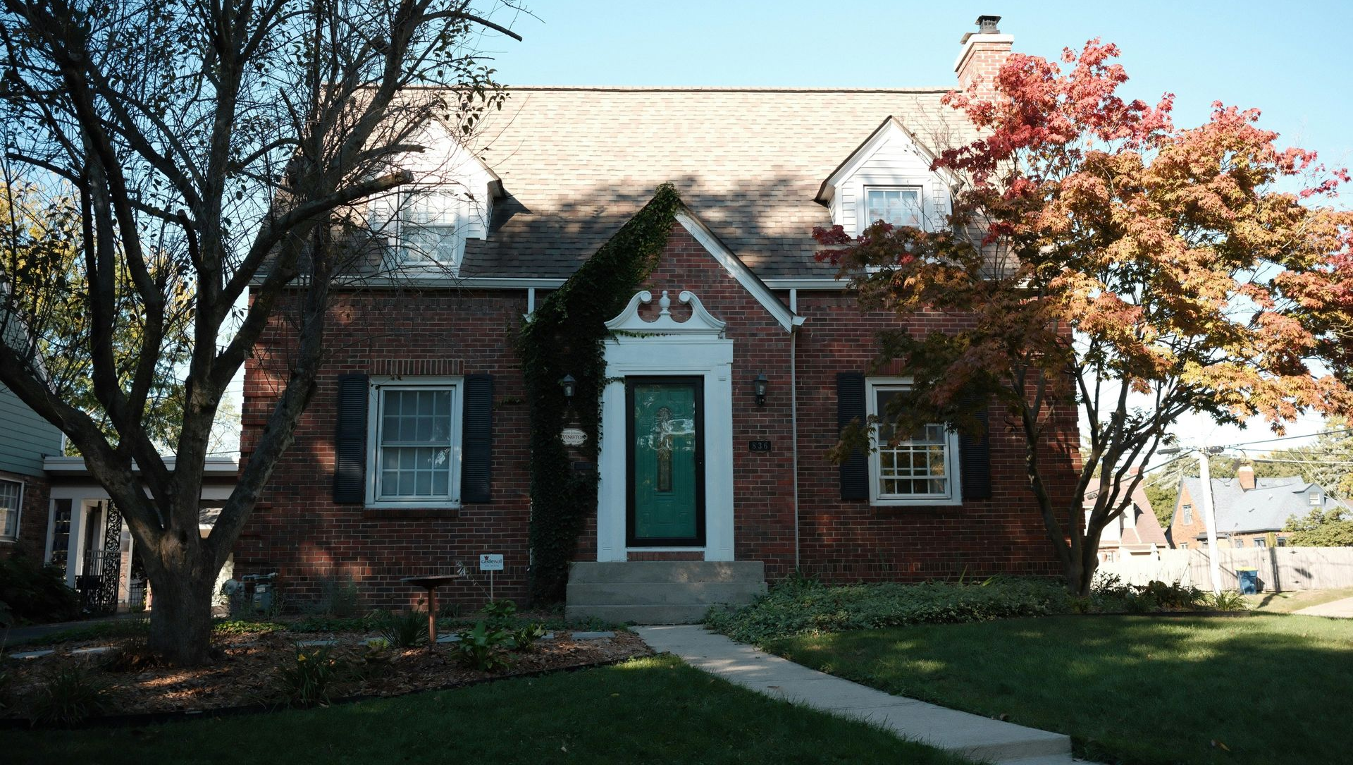 A one-story brick house with a steep roof, two dormer windows, and a central front door surrounded by autumn trees.