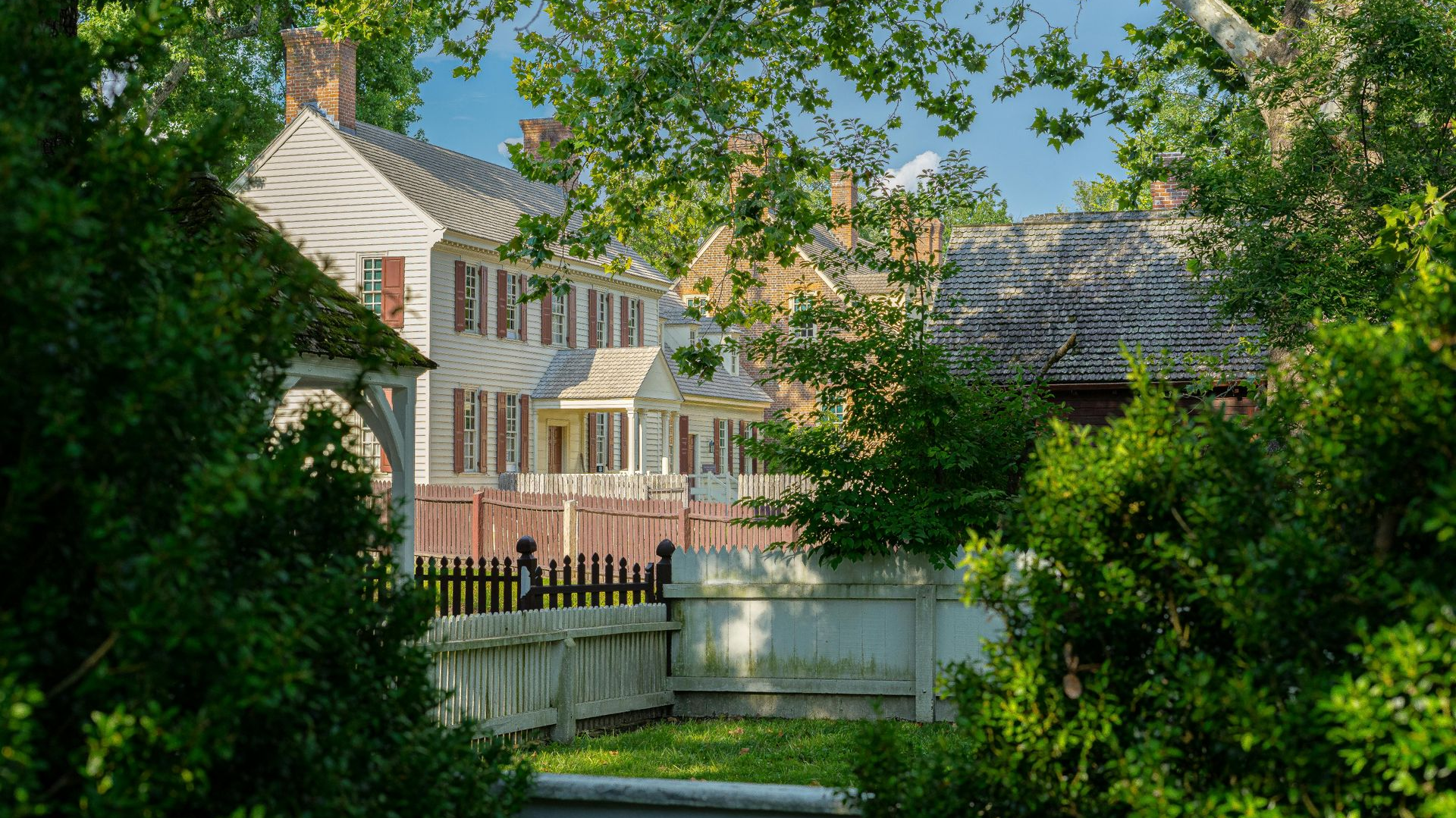 Historic homes with red shutters behind a white picket fence, framed by green foliage.