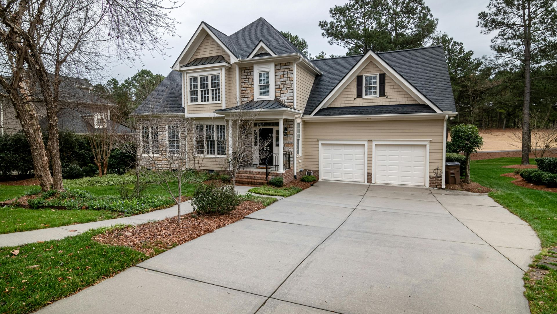 Two-story beige house with gray roof and stone accents, two-car garage, and concrete driveway.