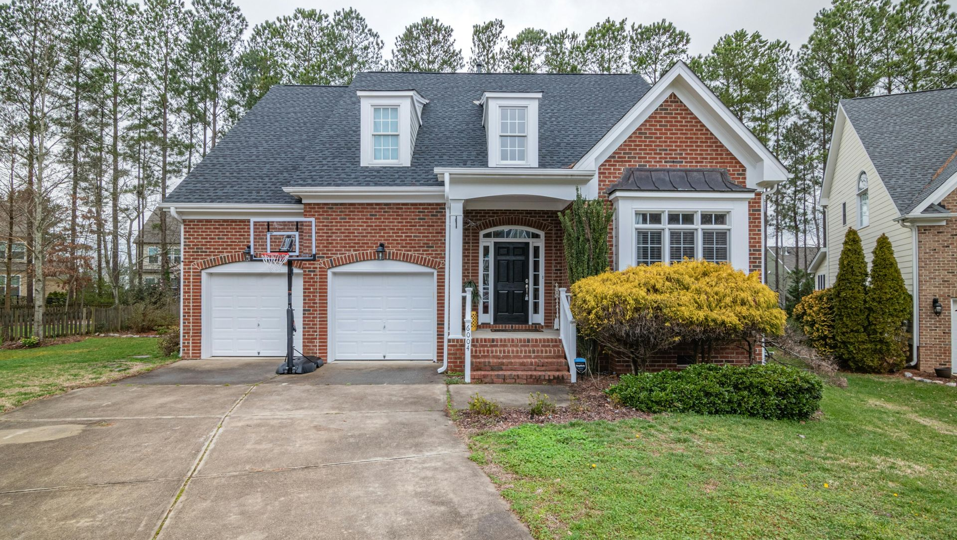A two-story red brick house with white trim, a two-car garage, and a small front porch, surrounded by trees and a lawn.