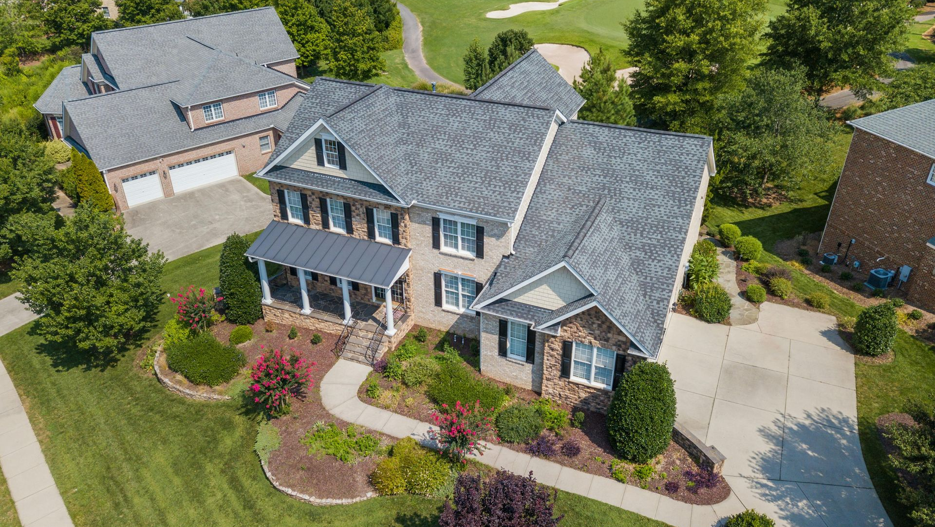 Aerial view of a large suburban brick house with a gray shingled roof, surrounded by green lawns and golf course grounds.