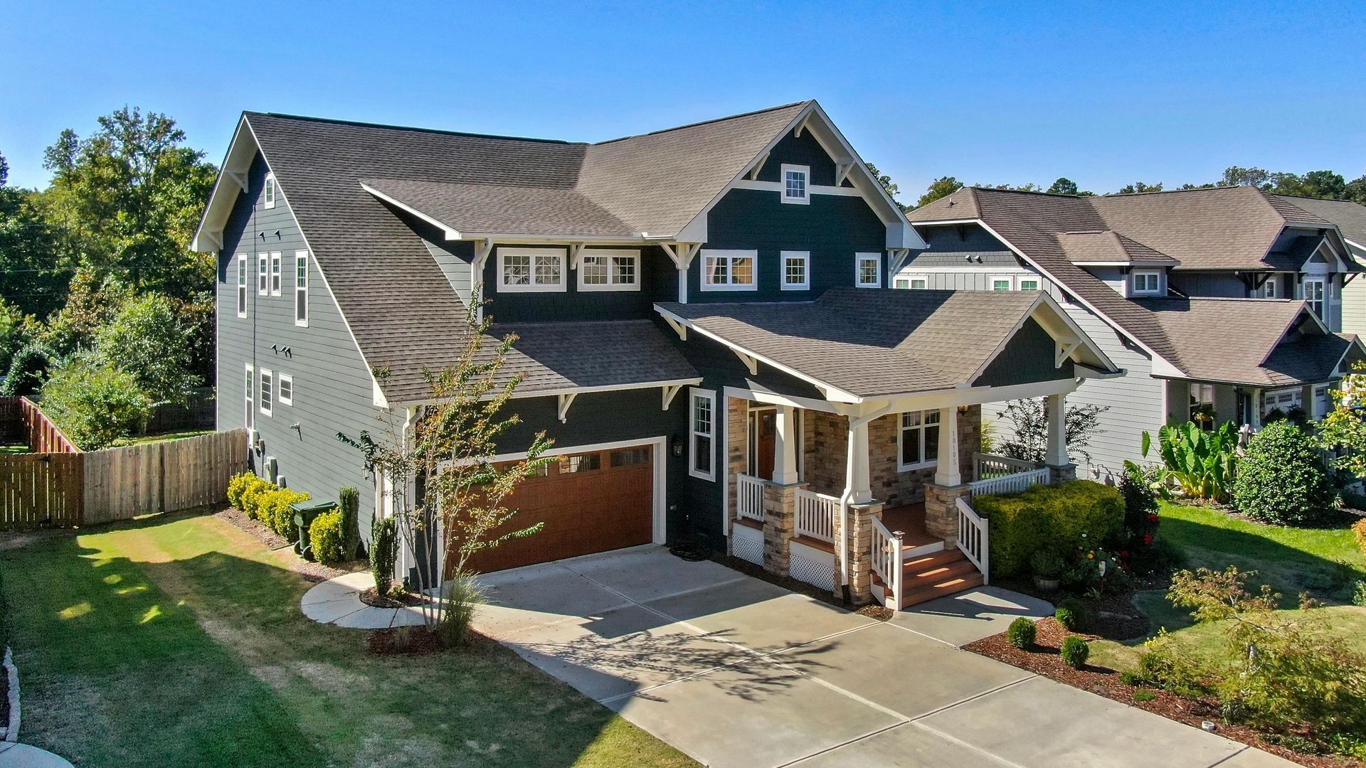 A two-story dark green home with a stone-accented porch, a garage, and a brown shingled roof under a clear blue sky.