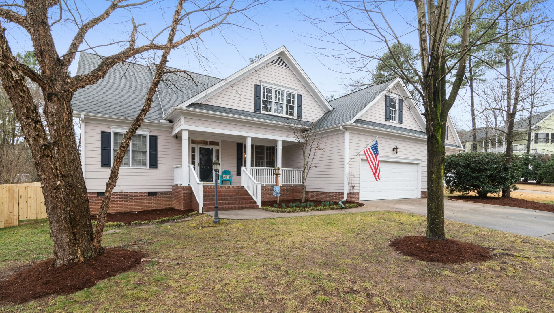 A light-colored two-story suburban house with a front porch, a brick foundation, and an American flag near the garage.