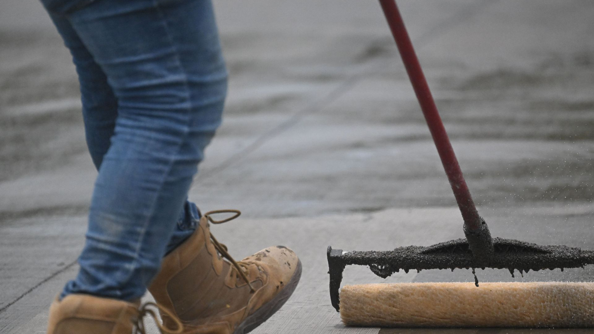 Worker pushing a black asphalt roller across wet pavement while wearing jeans and tan boots