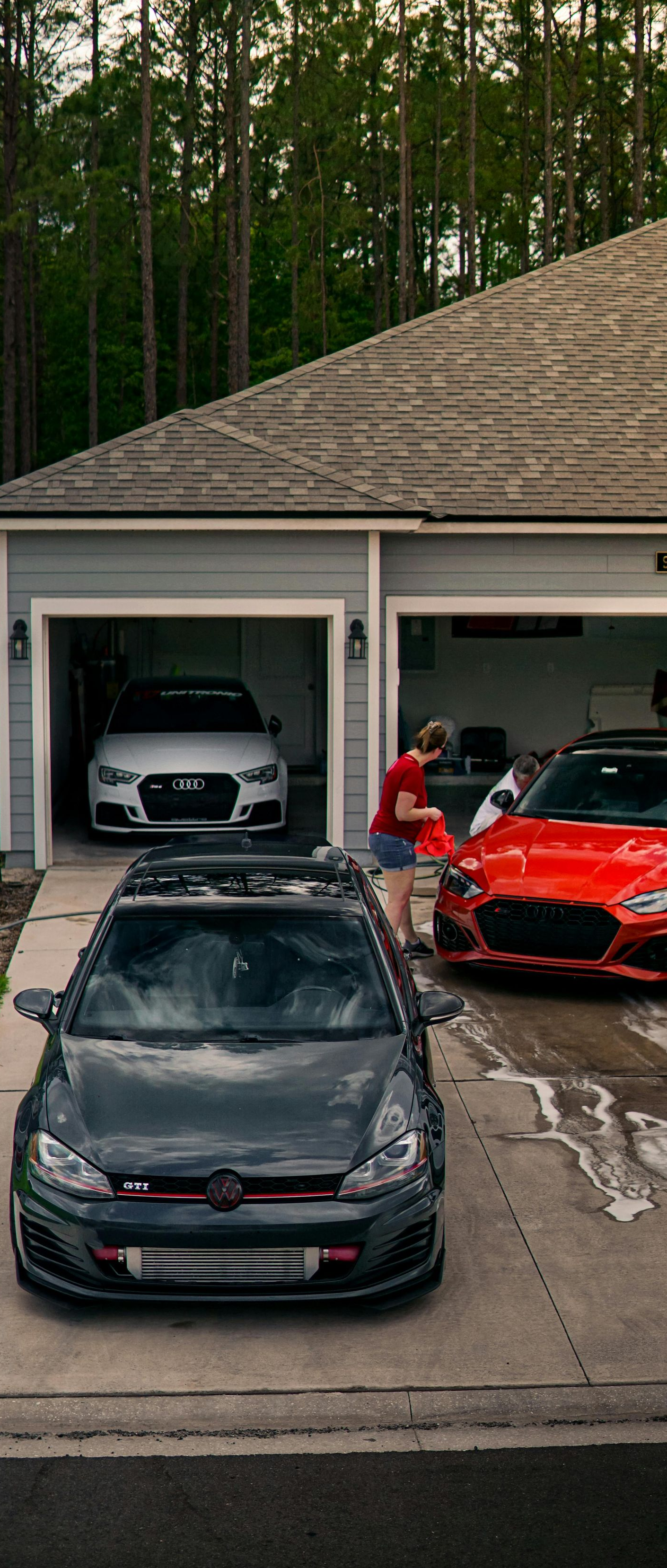 Two sports cars and a dark sedan parked in a driveway outside a garage, with trees behind the house.