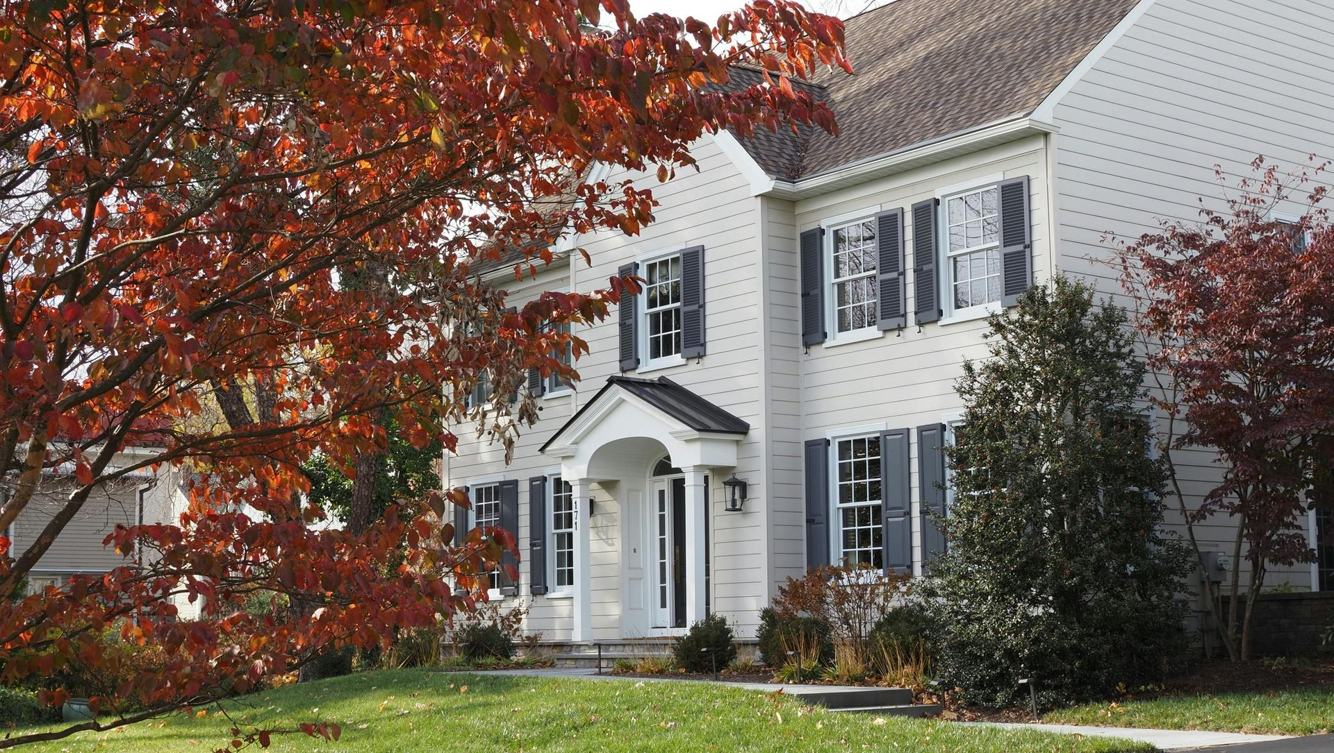 A two-story white house with dark shutters, centered in a suburban yard with autumn trees and a paved driveway.
