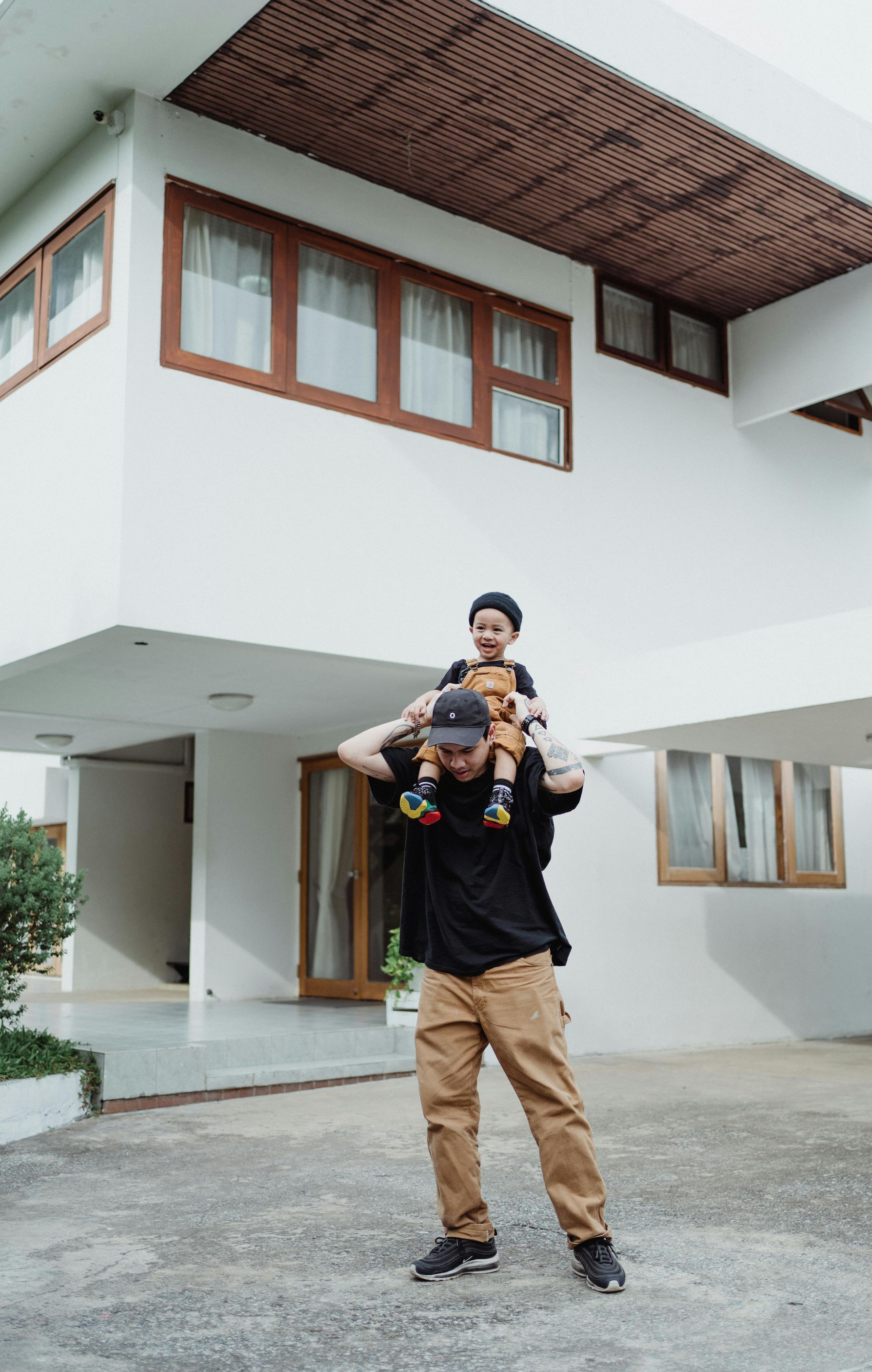 A person carries a child on their shoulders in front of a white two-story house with a wooden balcony and trim.