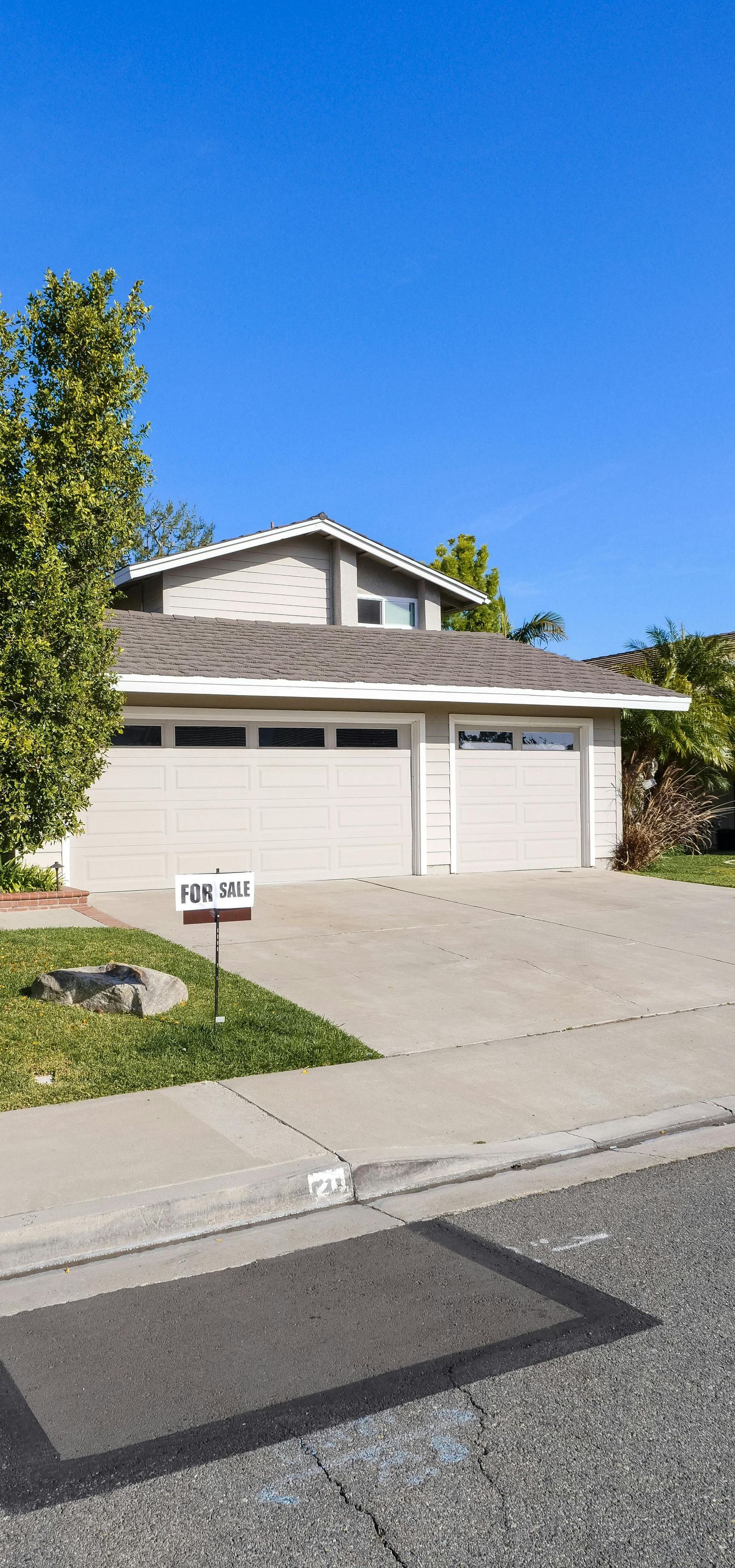 A beige suburban house with a two-car garage and a small tree in the front yard under a clear blue sky.
