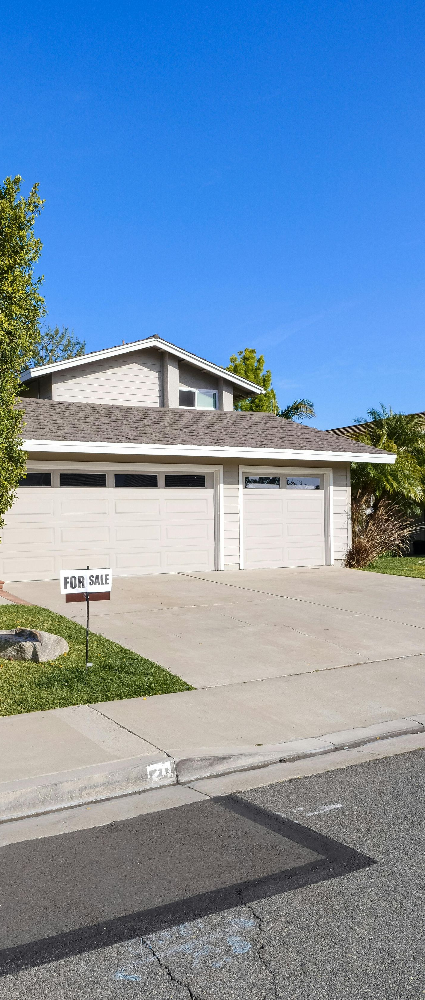 A light gray house with a two-car garage and a small tree in the front yard under a clear blue sky.