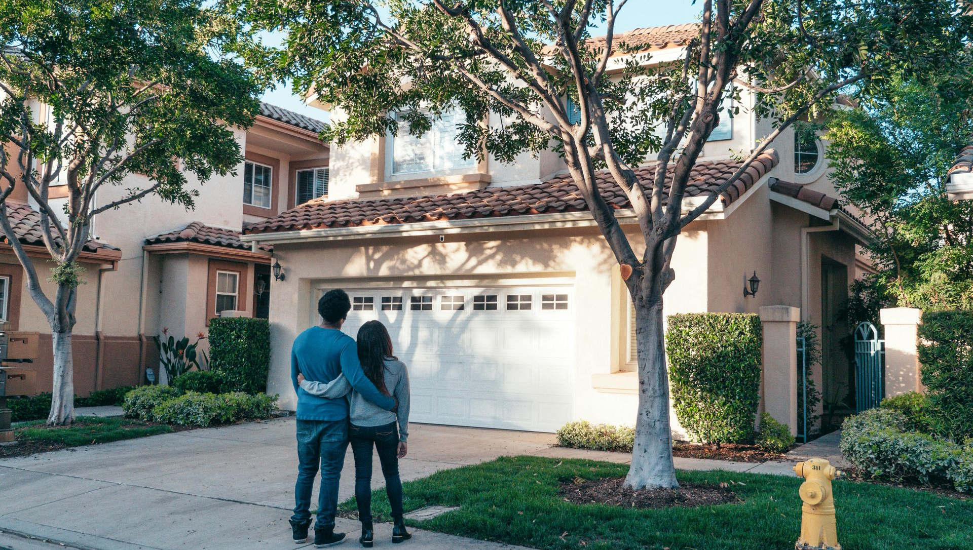 Couple embracing, looking at two-story home with white garage door, trees, and green lawn.