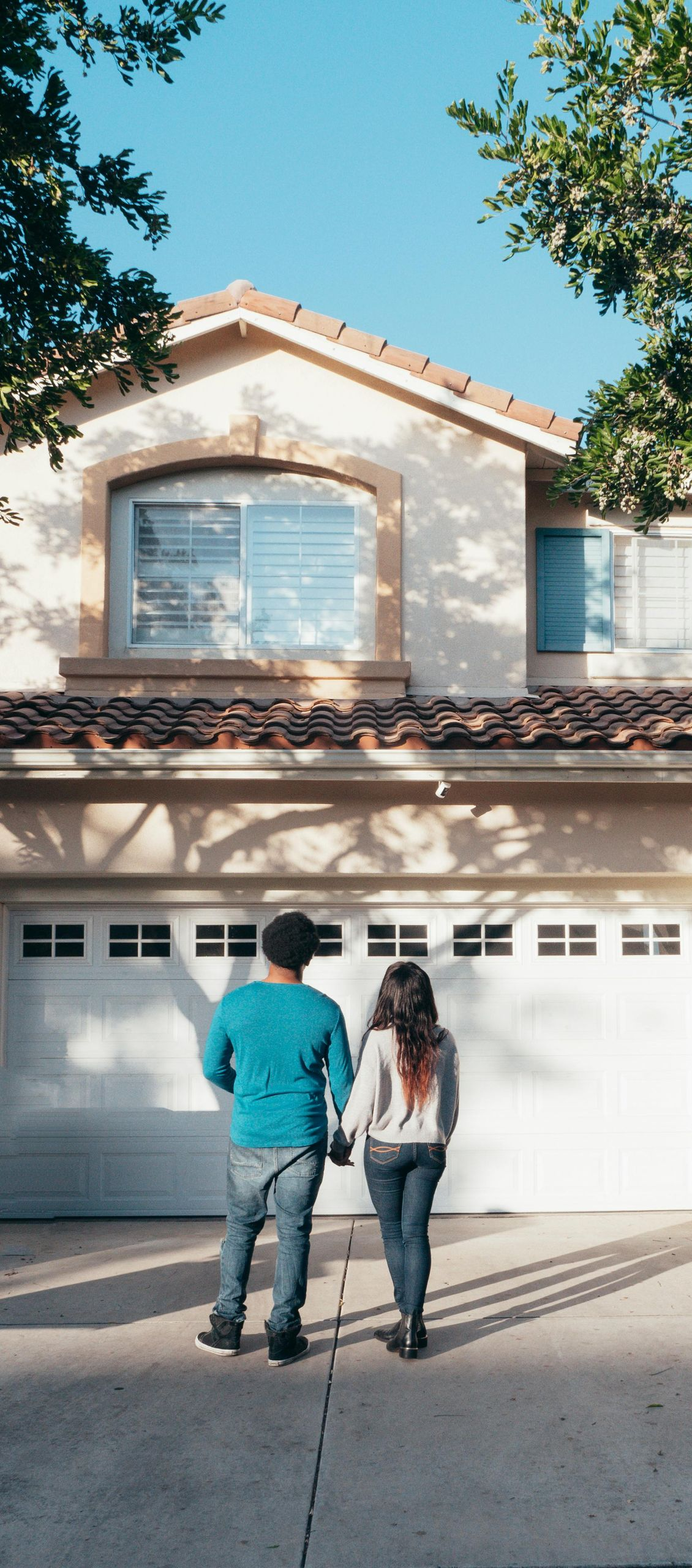A couple stands in a driveway, viewed from behind, looking toward a two-story beige house with a closed white garage.