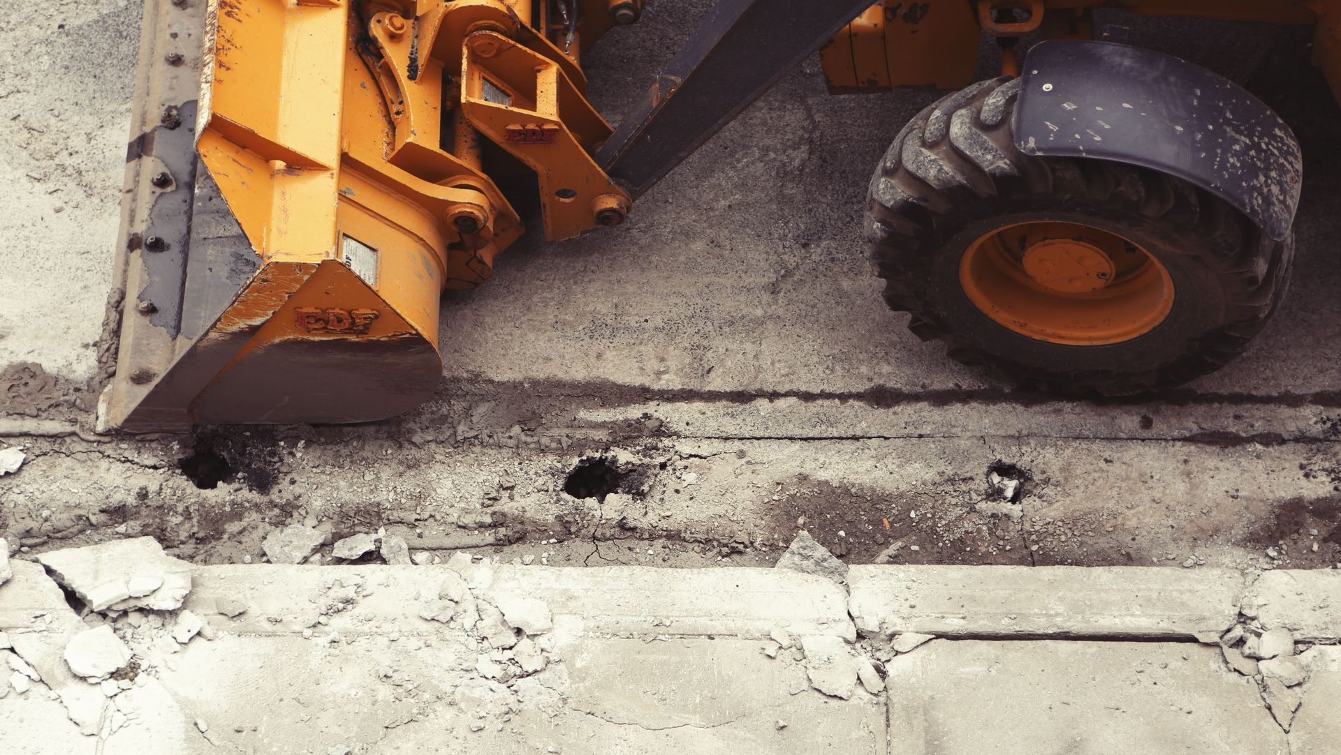Orange construction equipment resurfacing a broken concrete road at a curb edge