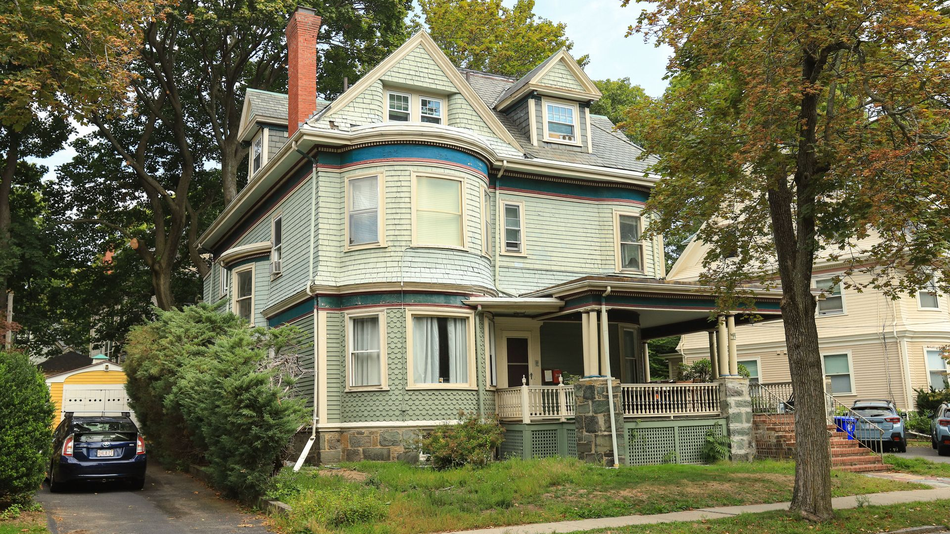 Old blue-gray Victorian house with a porch, trees, and parked cars along a residential street
