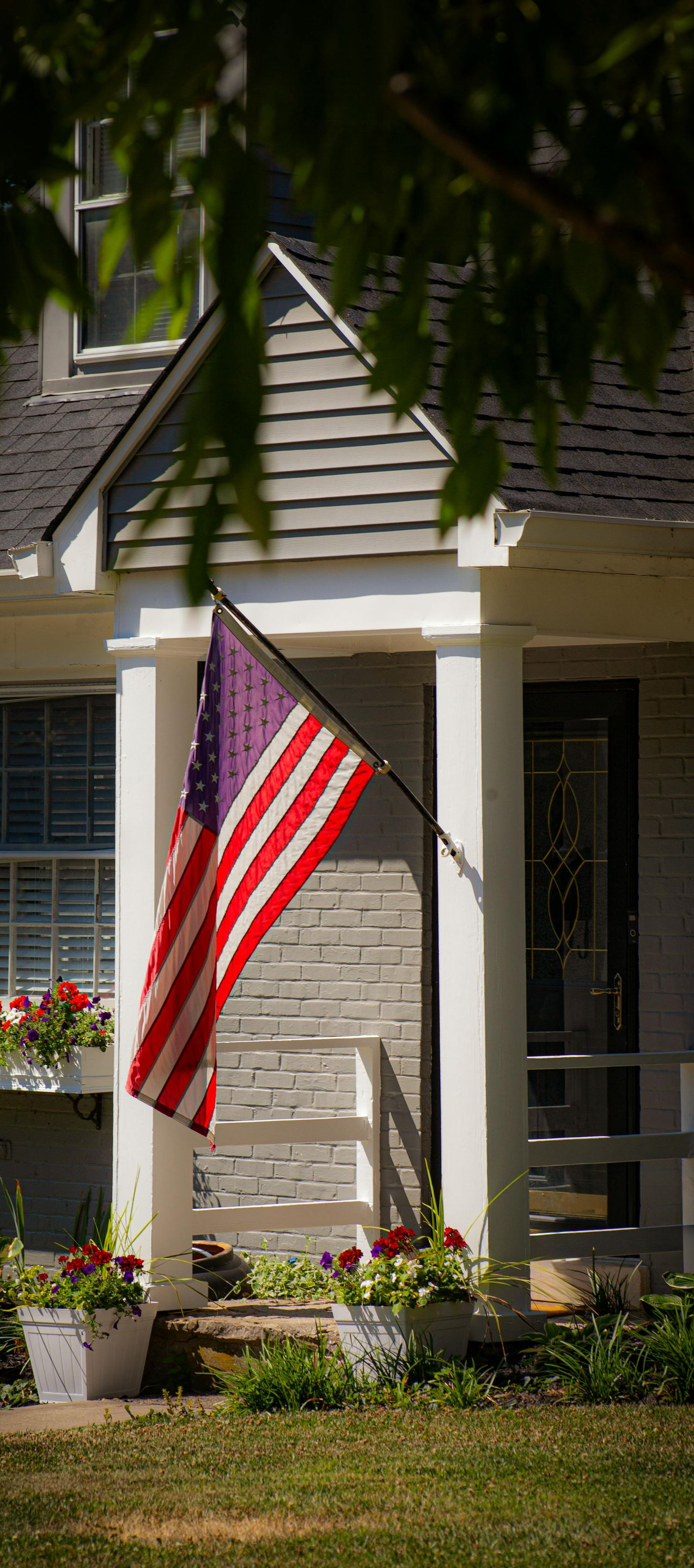 An American flag hangs from the front porch of a brick home with white columns and potted flowers.