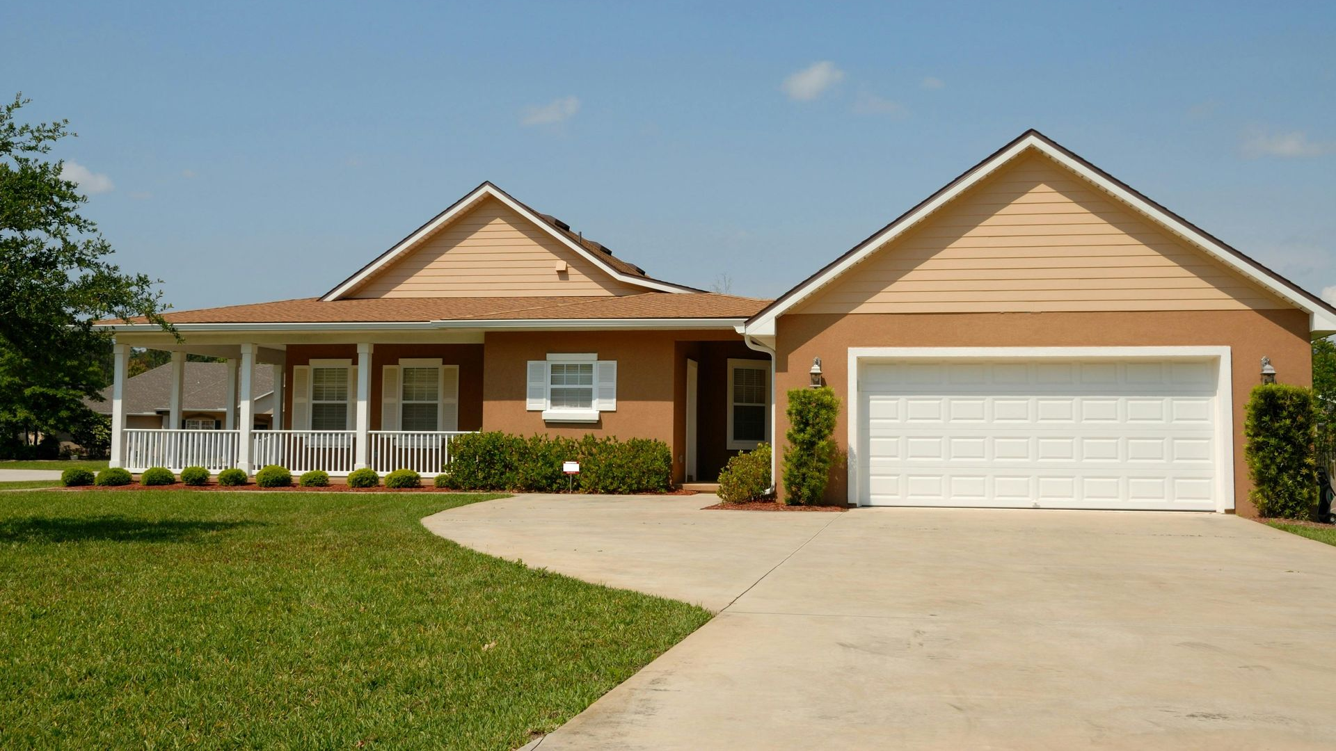 Tan-colored house with a porch and attached garage on a sunny day. Green lawn and blue sky.