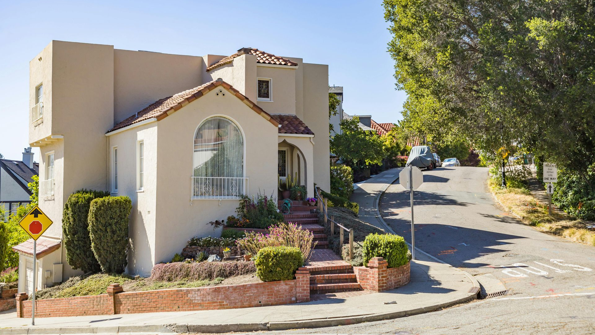Cream stucco house on a sunny hillside street with a large tree and brick steps