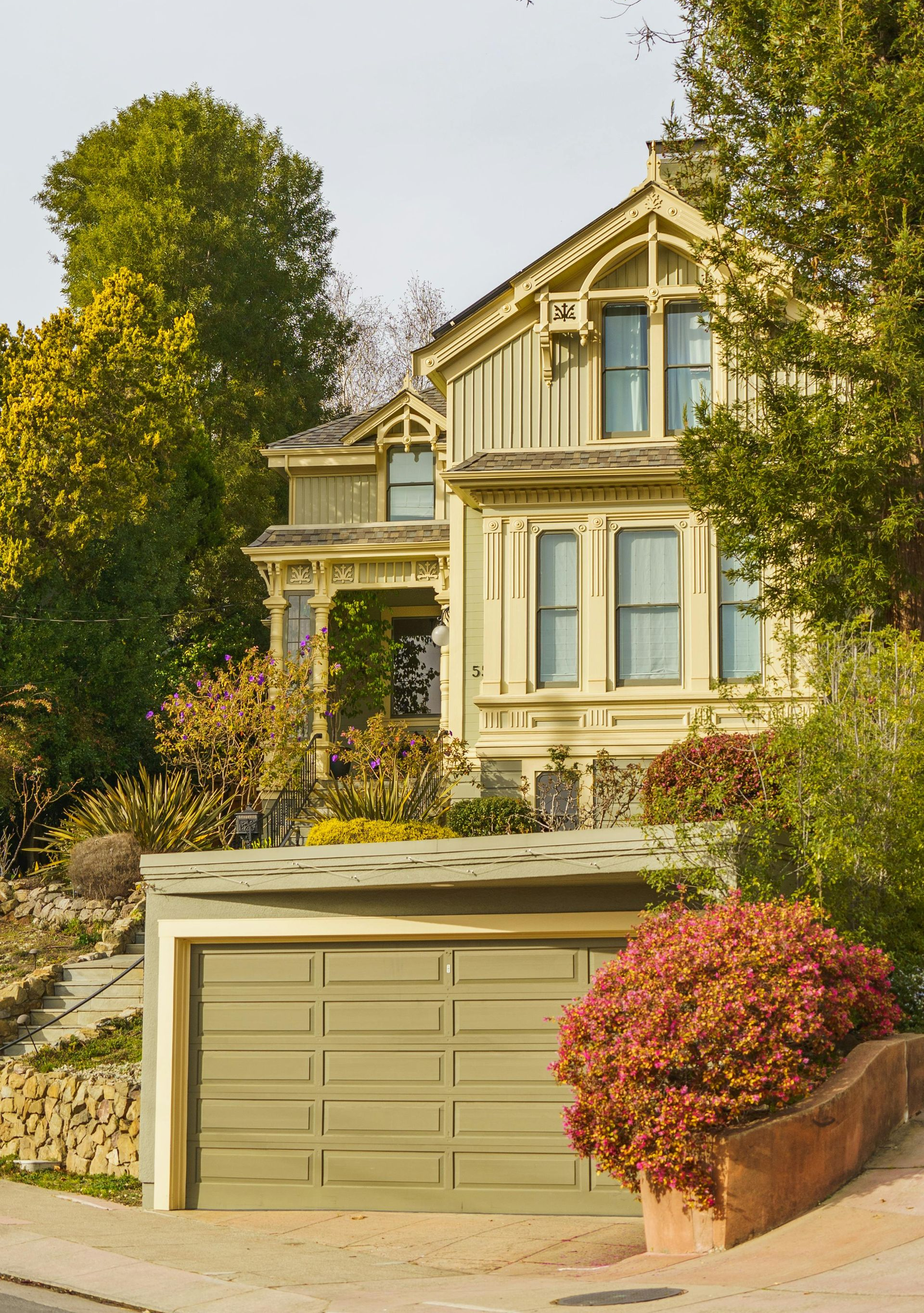A historic, pale yellow Victorian home with ornate trim, partially obscured by trees, above a detached garage.