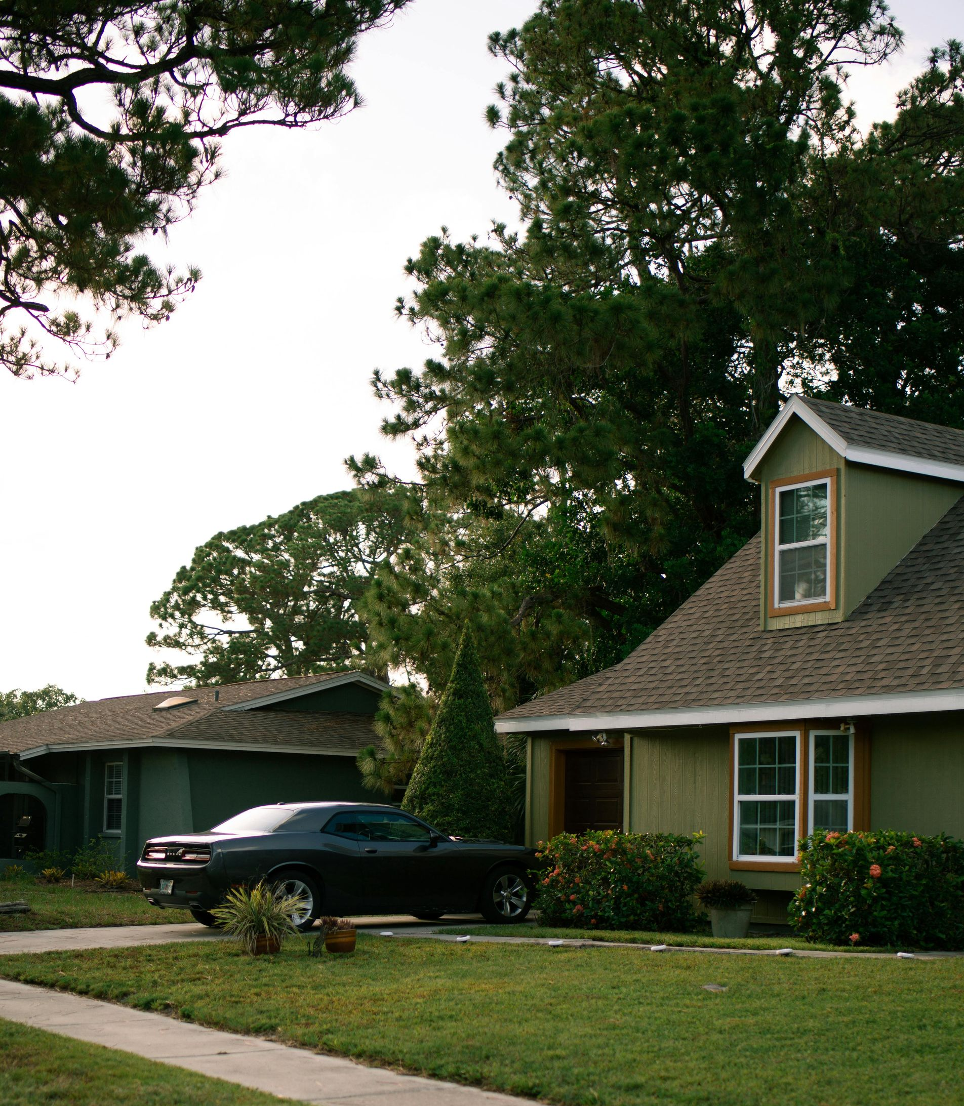 A black car parked in the driveway of a green cottage-style house with a front lawn and a sidewalk.