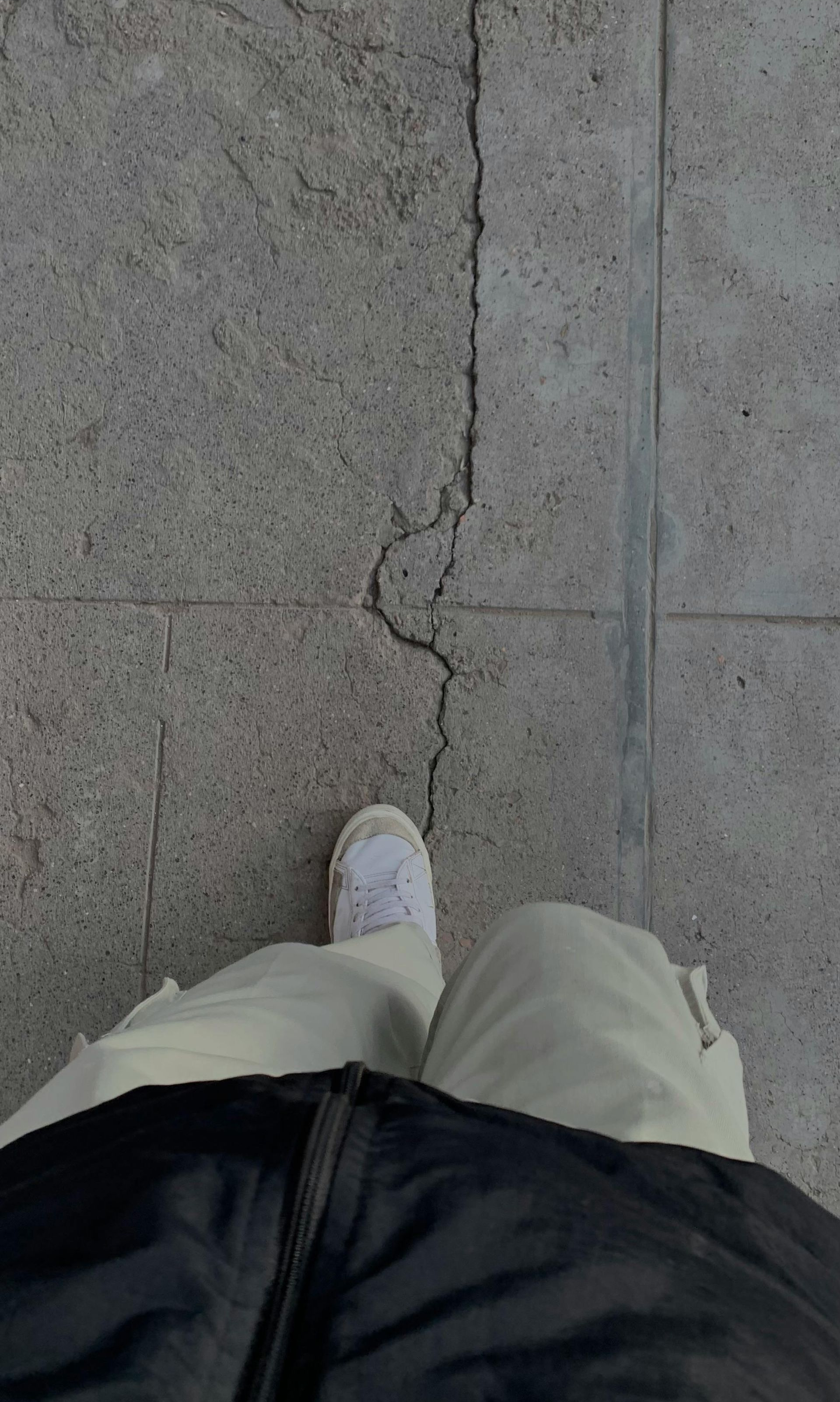 Person's feet on cracked pavement. White sneakers, beige pants, and black jacket visible.