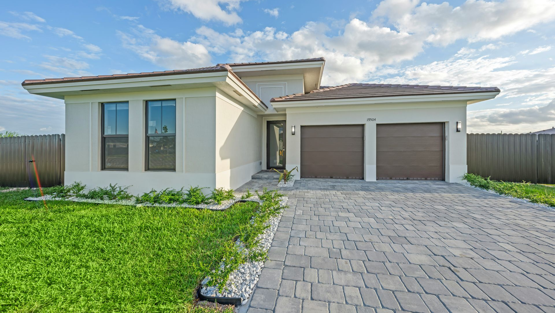 Modern beige house with a two-car garage, brick driveway, and green lawn under a partly cloudy sky.
