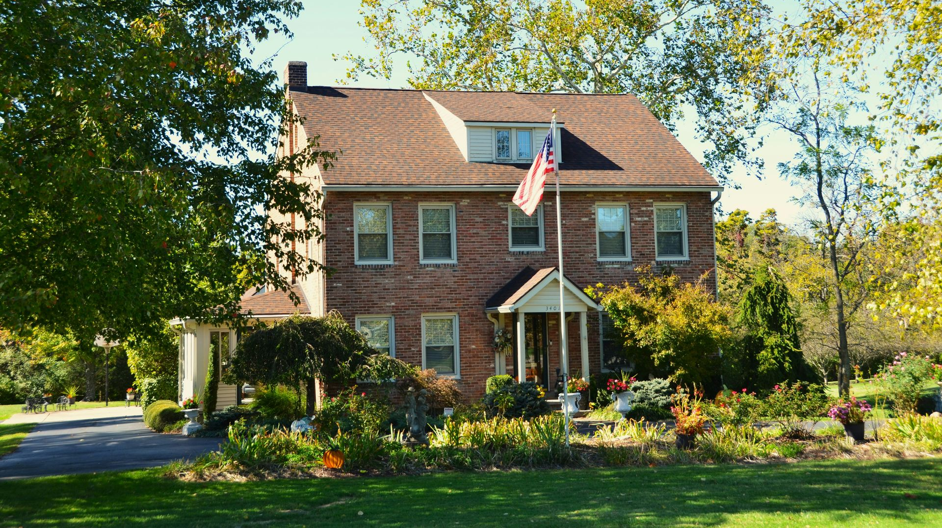 Two-story brick house with flag, trees, and garden on a sunny day.