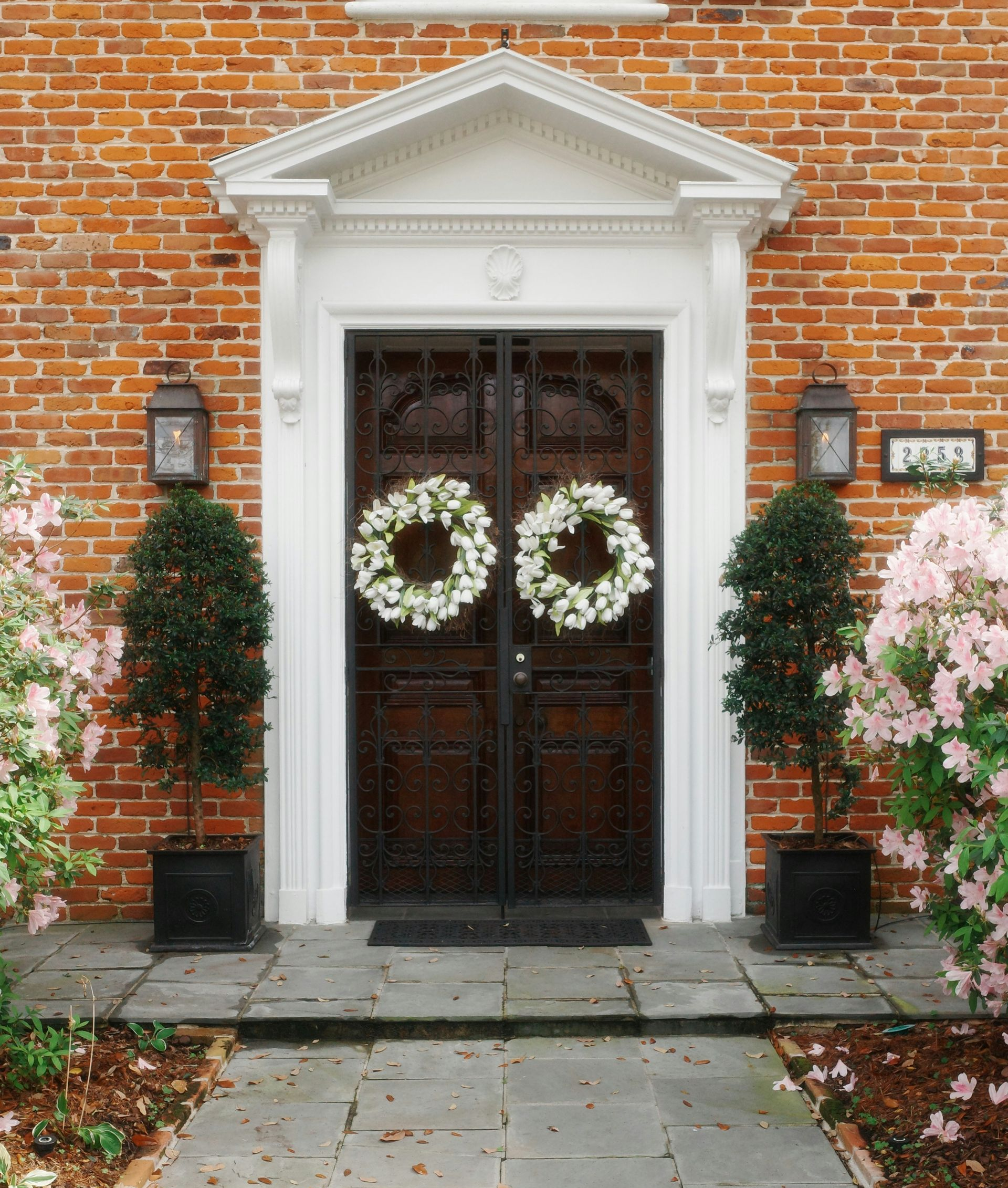A dark wooden double door with two white wreaths, framed by white molding, flanked by potted trees and brick walls.