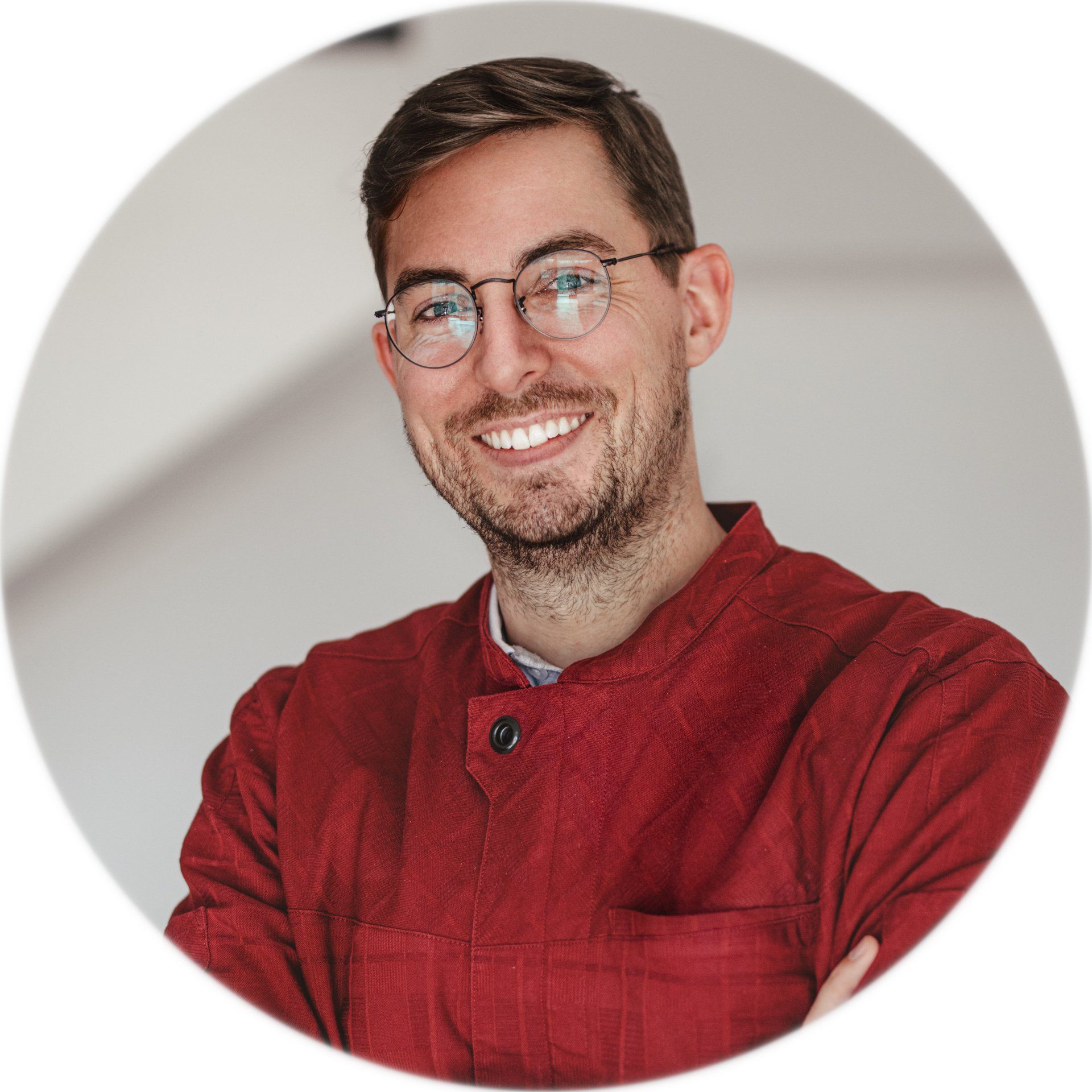Lächelnder Mann mit Brille, der eine rote Jacke trägt, die Arme verschränkt, vor hellem Hintergrund.