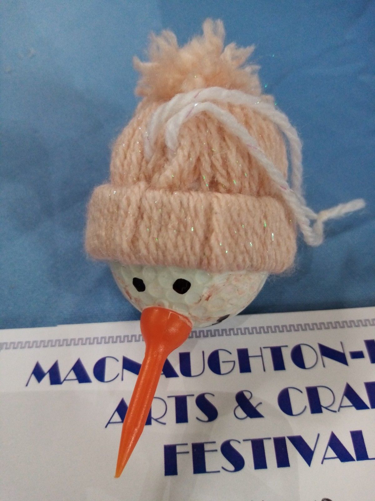 A snowman with a hat and a carrot stick is sitting on a sign that says macnaughton arts & craft festival