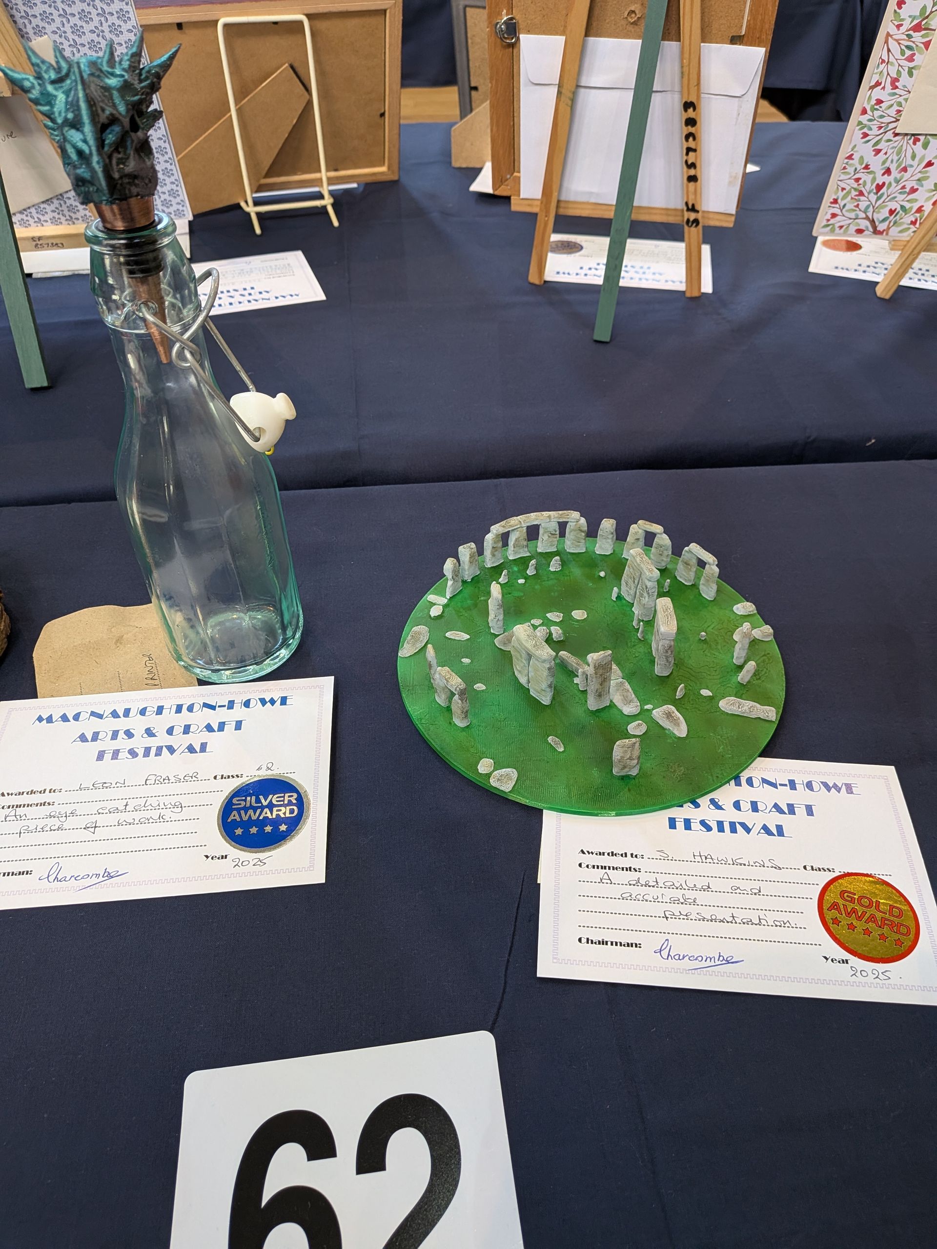 A table display includes a model of Stonehenge, a decorated bottle, and number 62.