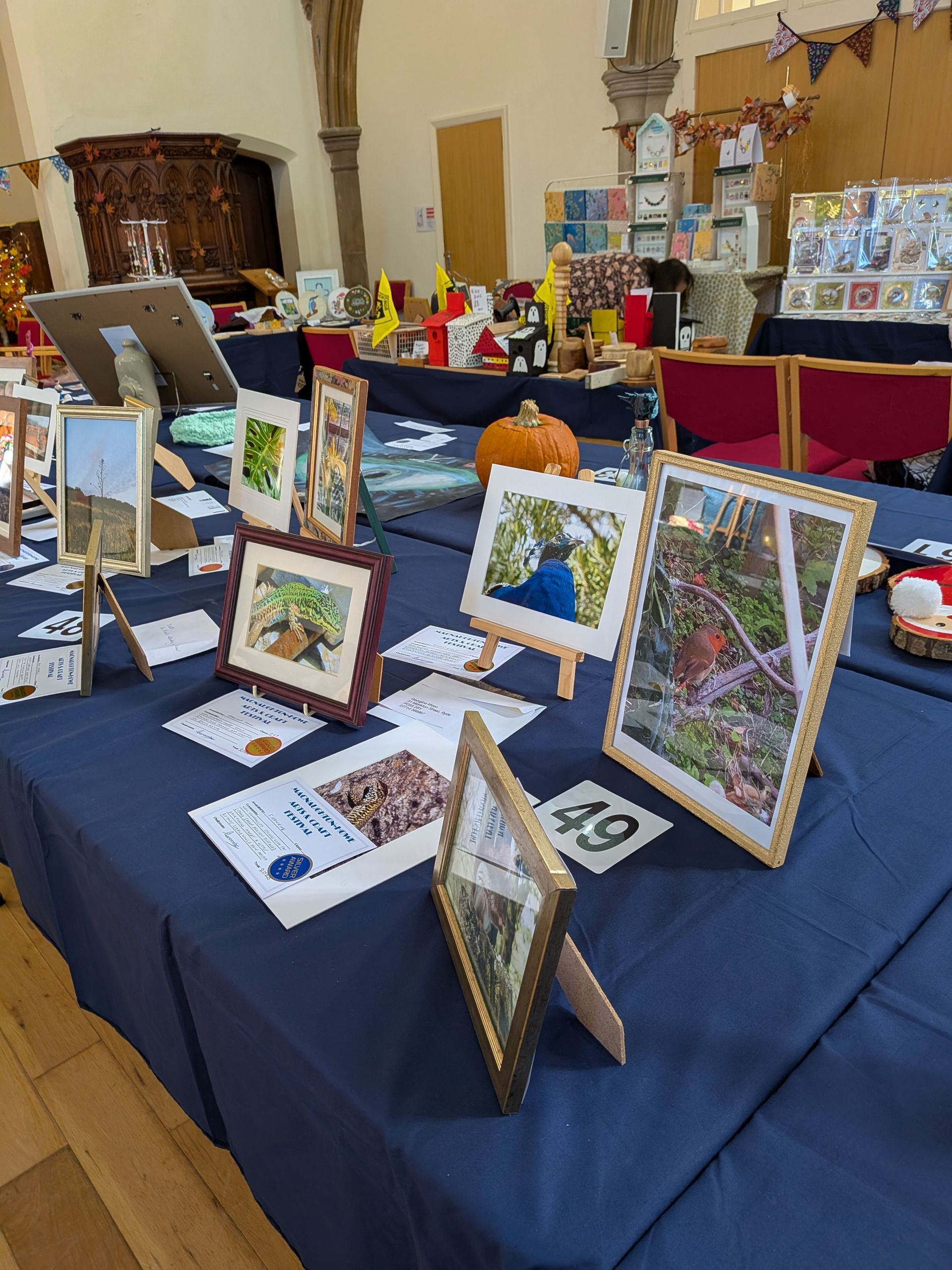 A display table at an art fair, featuring framed artwork, cards, and a pumpkin.