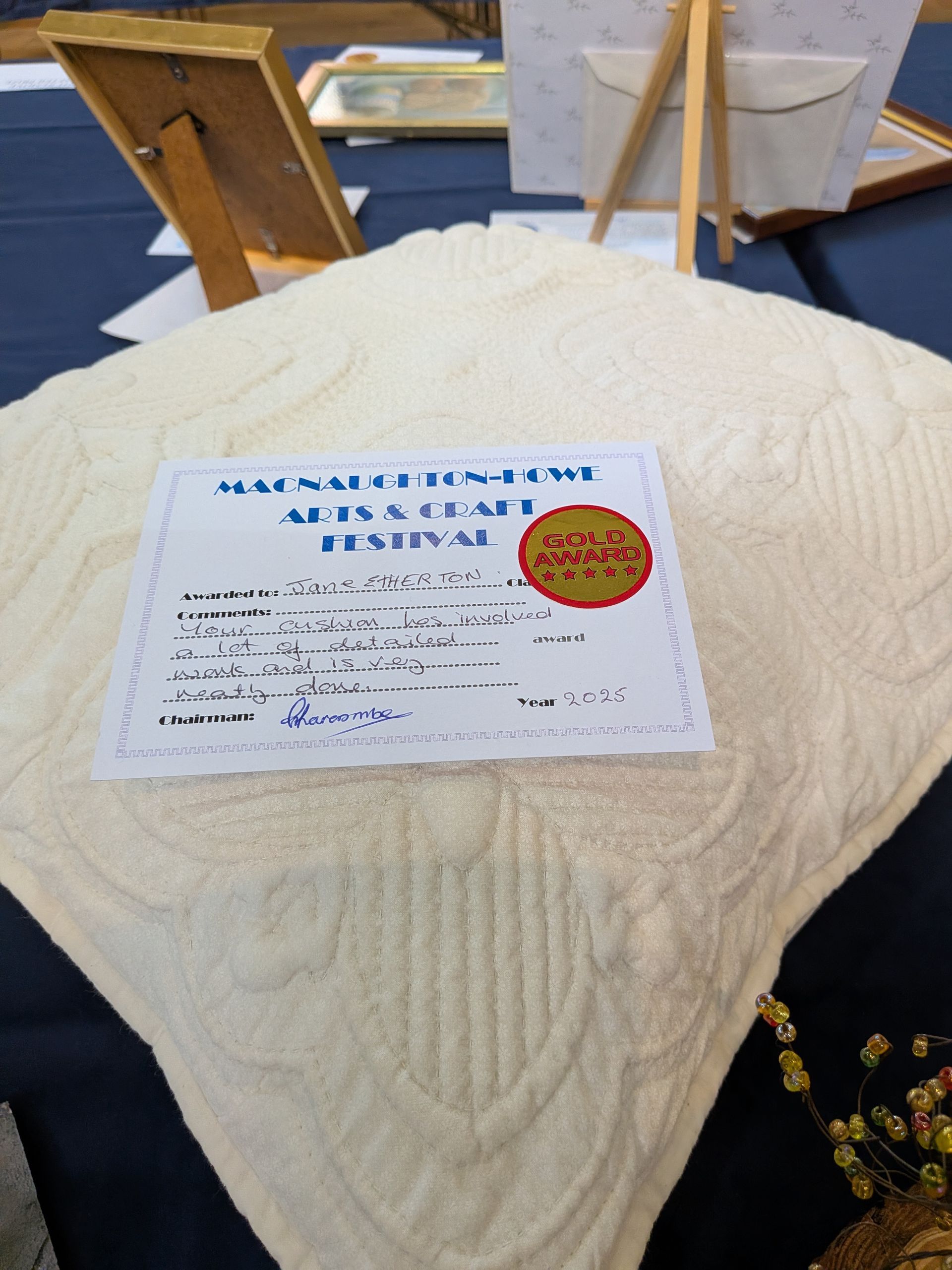 Cream-colored quilt with a festival award certificate on a blue tablecloth. Wooden easels and decor are in the background.