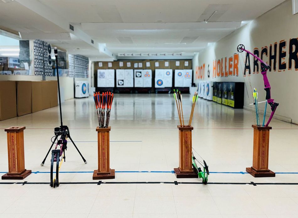 Indoor archery range with bows on stands, target lanes, and a tripod in the foreground.