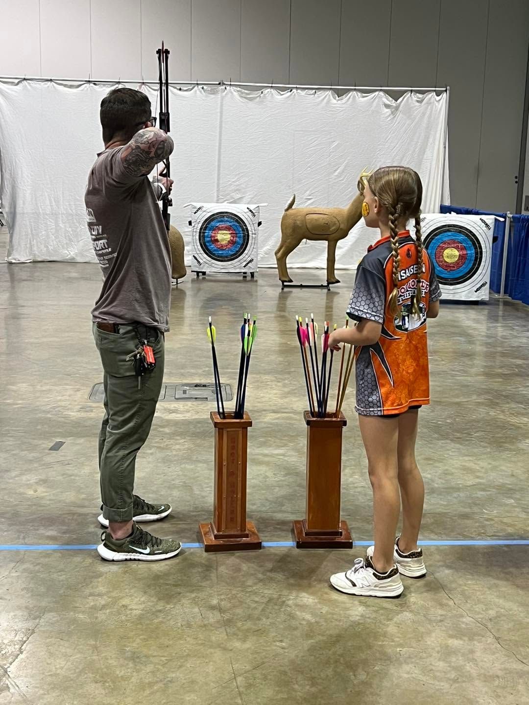Indoor archery range with two people aiming at targets, plus a deer target in the background.