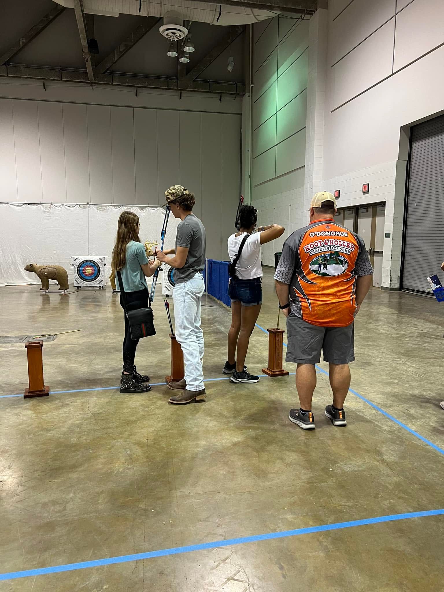 People standing in an indoor hall, some aiming pistols at targets during a shooting event.