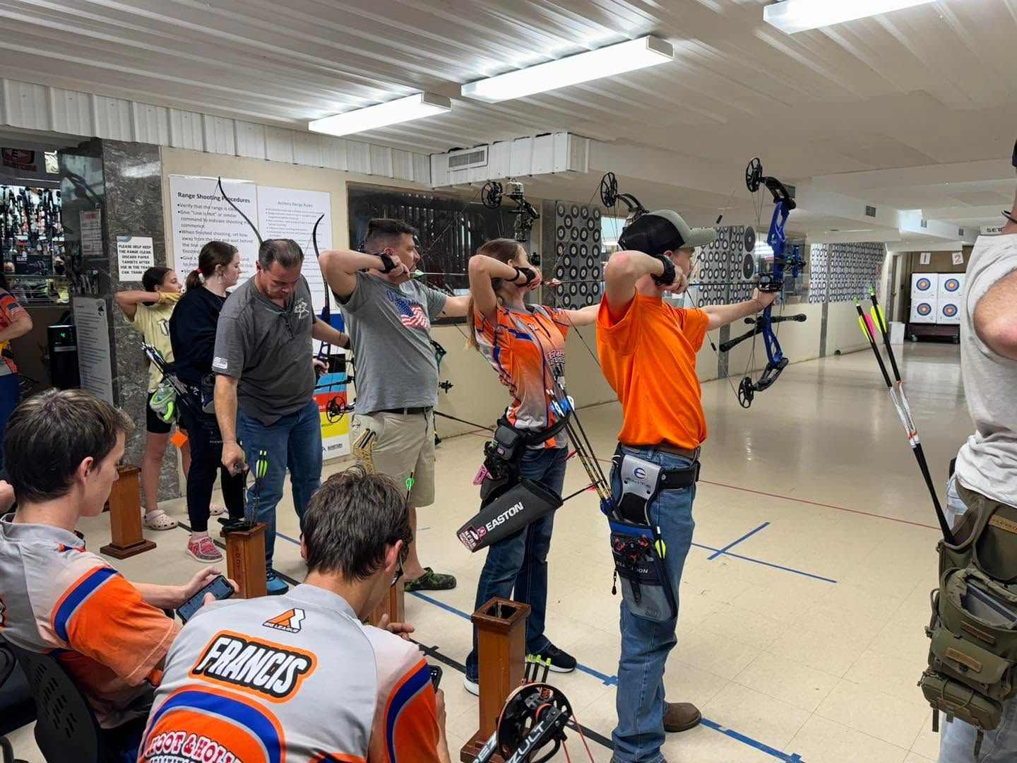 Archery class indoors, with people in orange vests aiming bows at targets.