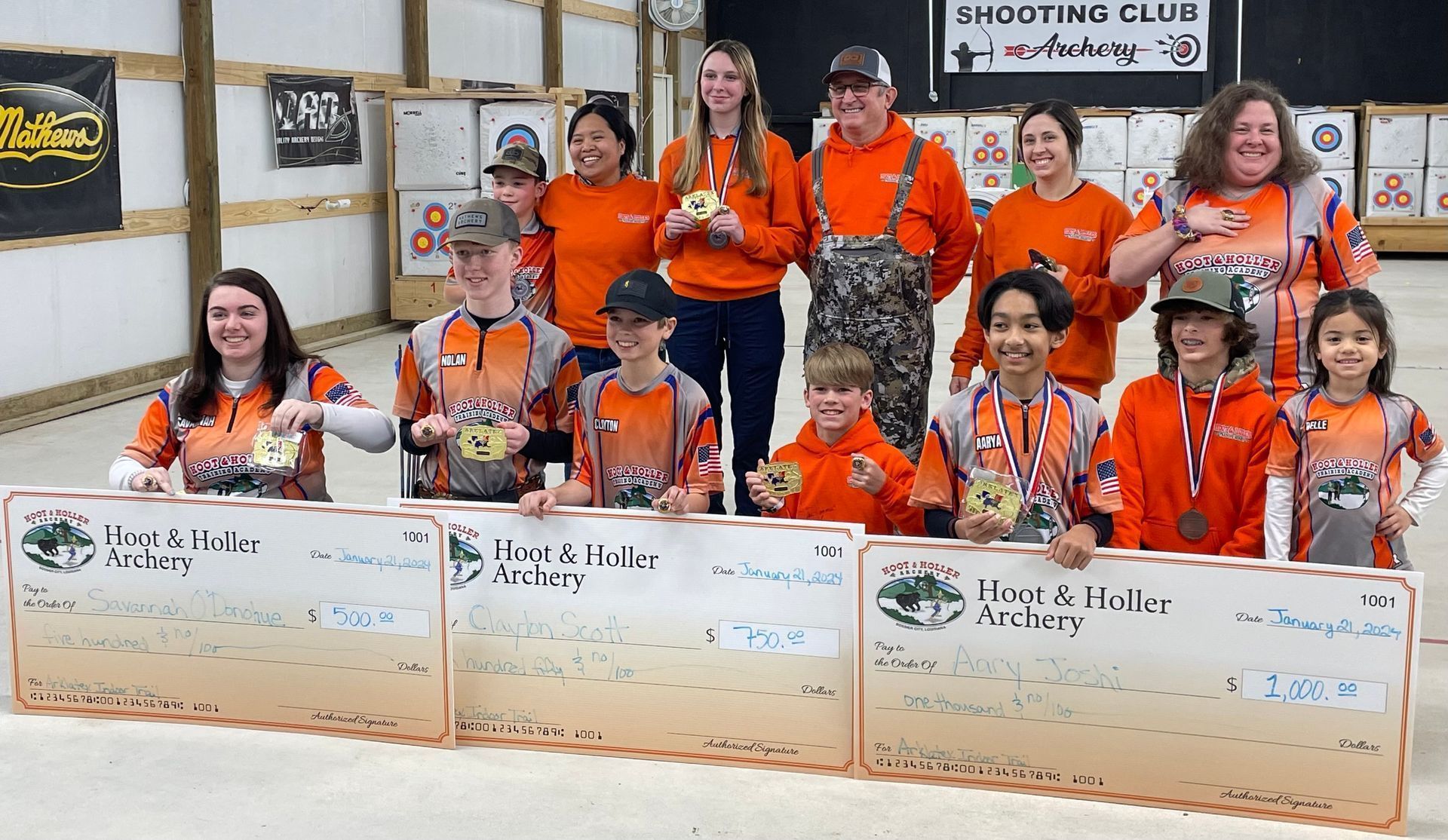 Group in orange shirts posing with certificates and large check boards at a skating club event