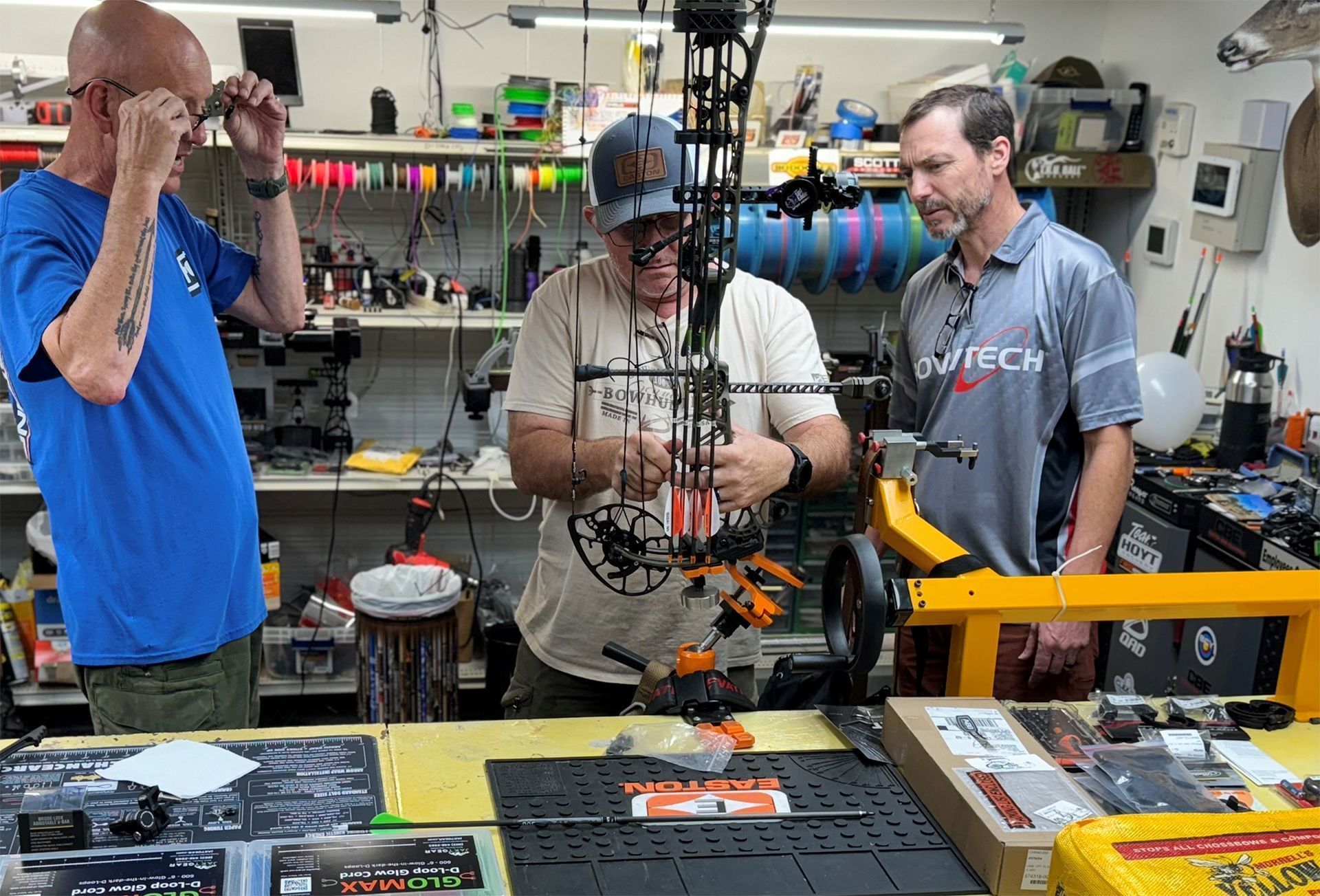 Three men in a workshop inspect a compound bow on a table.