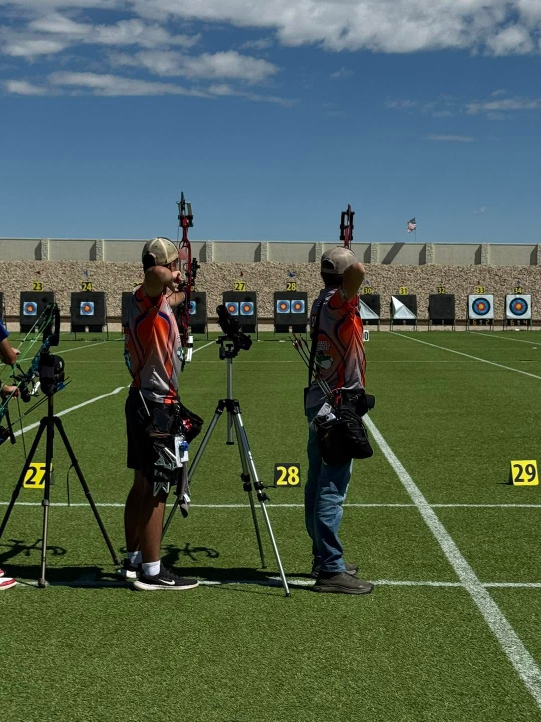 Two archers in orange shirts and hats stand on a field beside numbered shooting lanes and targets.